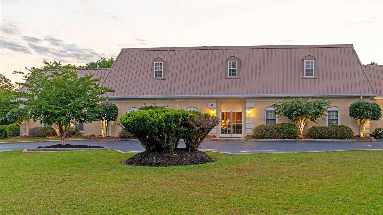 A beige building with a brown roof and arched windows sits on a green lawn with a large bush in the foreground.
