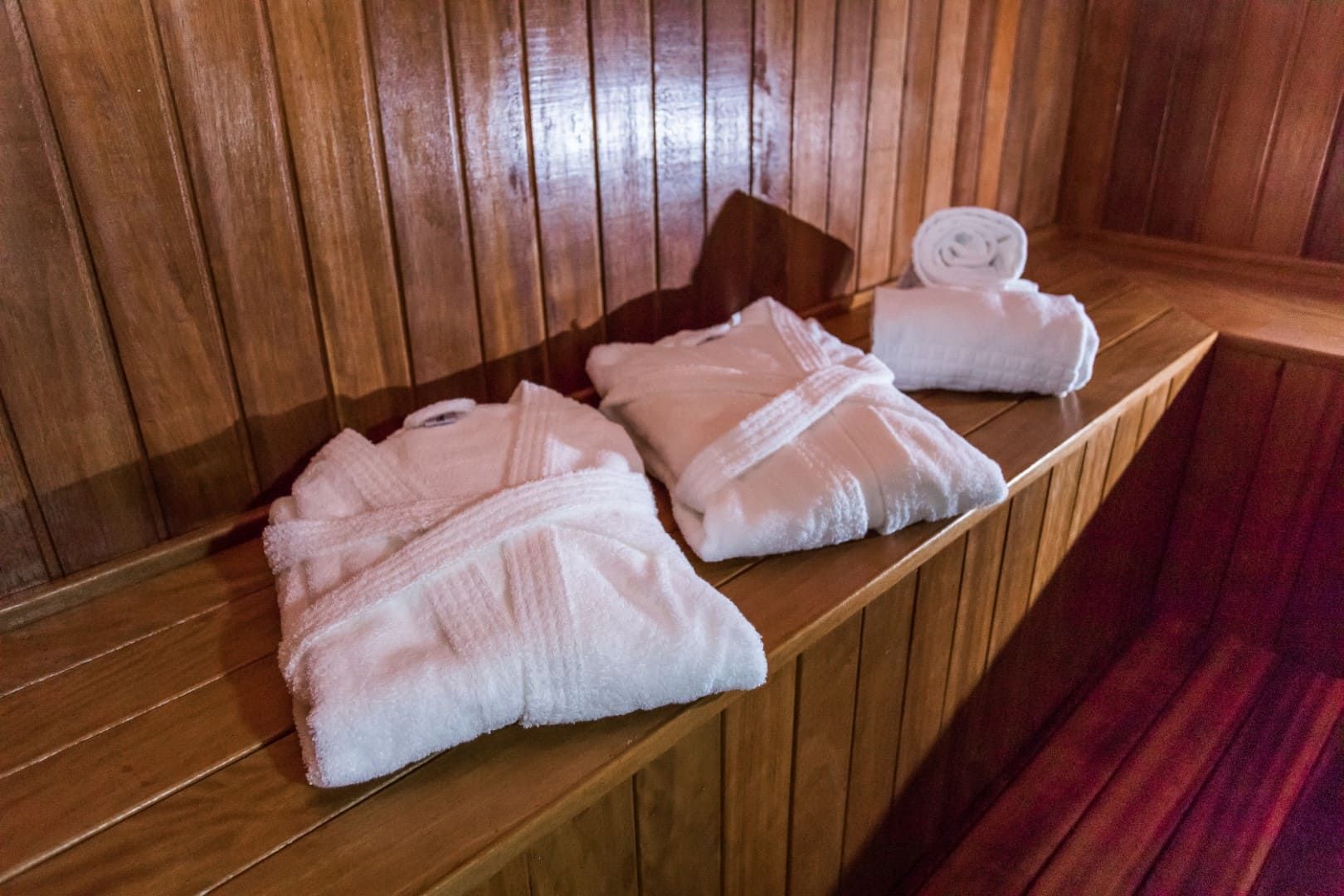 Dois roupões de banho e duas toalhas estão sobre um banco de madeira em uma sauna.