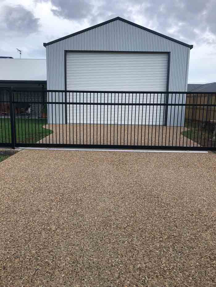 Black Gate and Gravel Driveway Leading to A Metal Shed with A Closed Garage Door — Wieland Concreting in Rockhampton, QLD