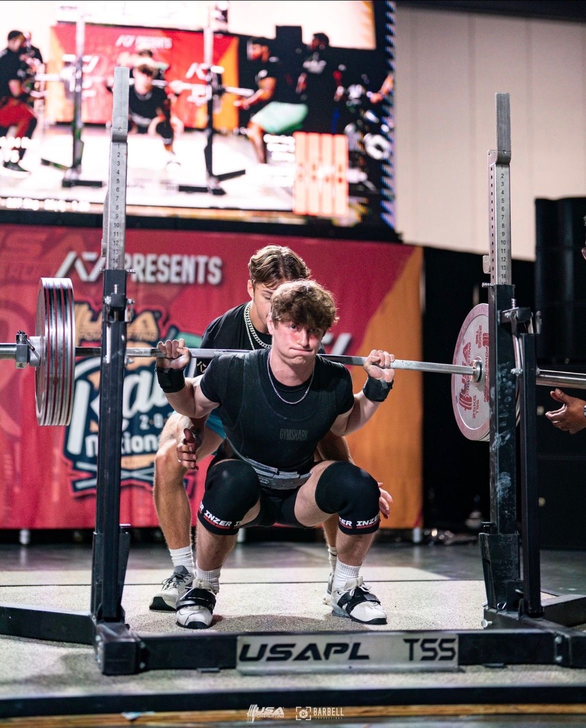 A powerlifter performs a barbell squat in a rack with a spotter standing behind, set against a competition stage backdrop.