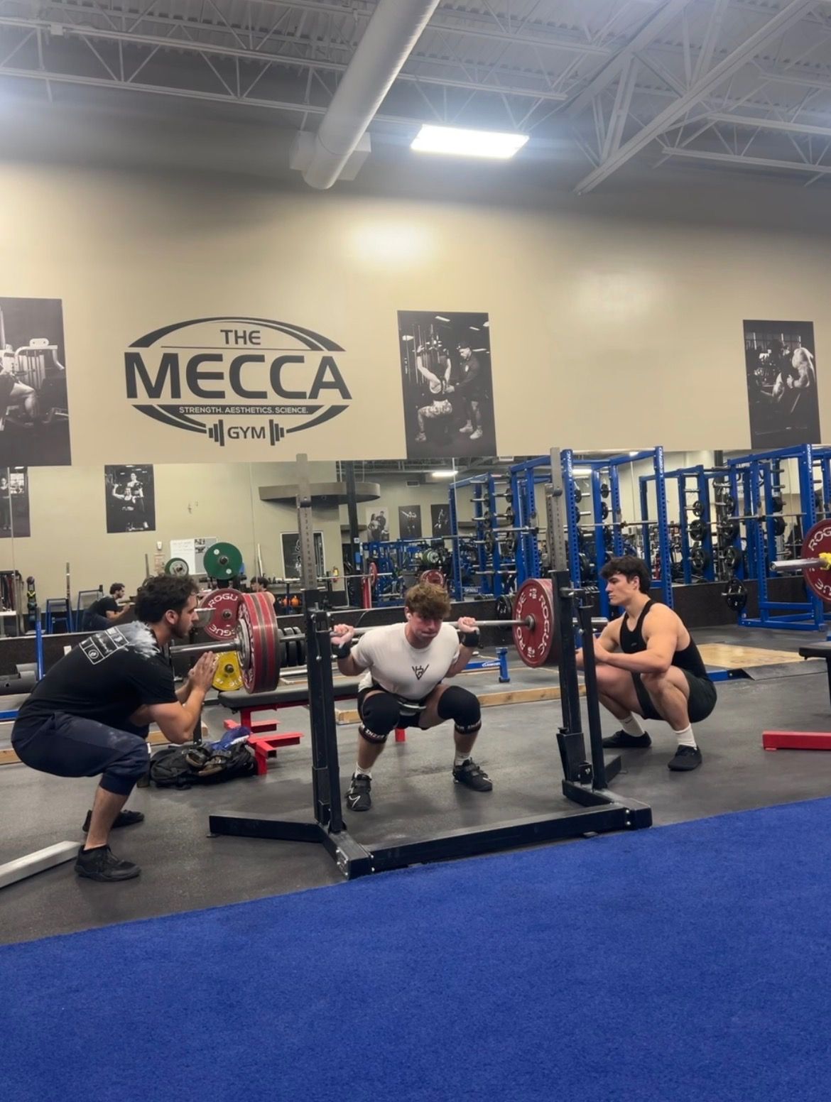 Three people in a gym perform a barbell squat with two individuals acting as spotters at a facility named The Mecca.