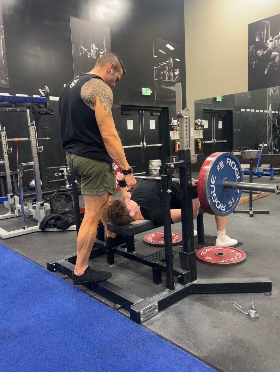 A person spotting another as they perform a bench press on a weight rack in a gym.