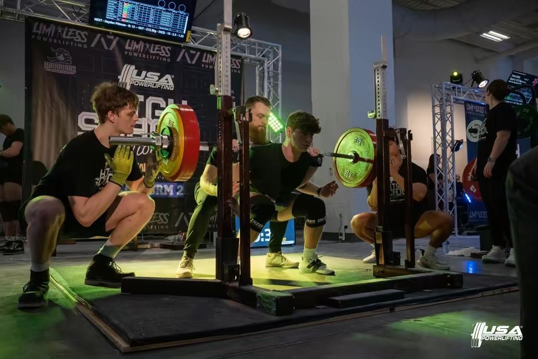 Three weightlifters performing a coordinated squat lift during an indoor competition.