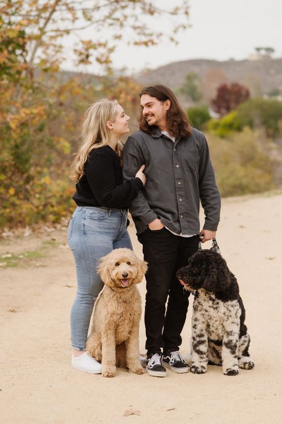 A man and a woman are standing next to two dogs on a dirt road. Orange county hair and makeup artist.