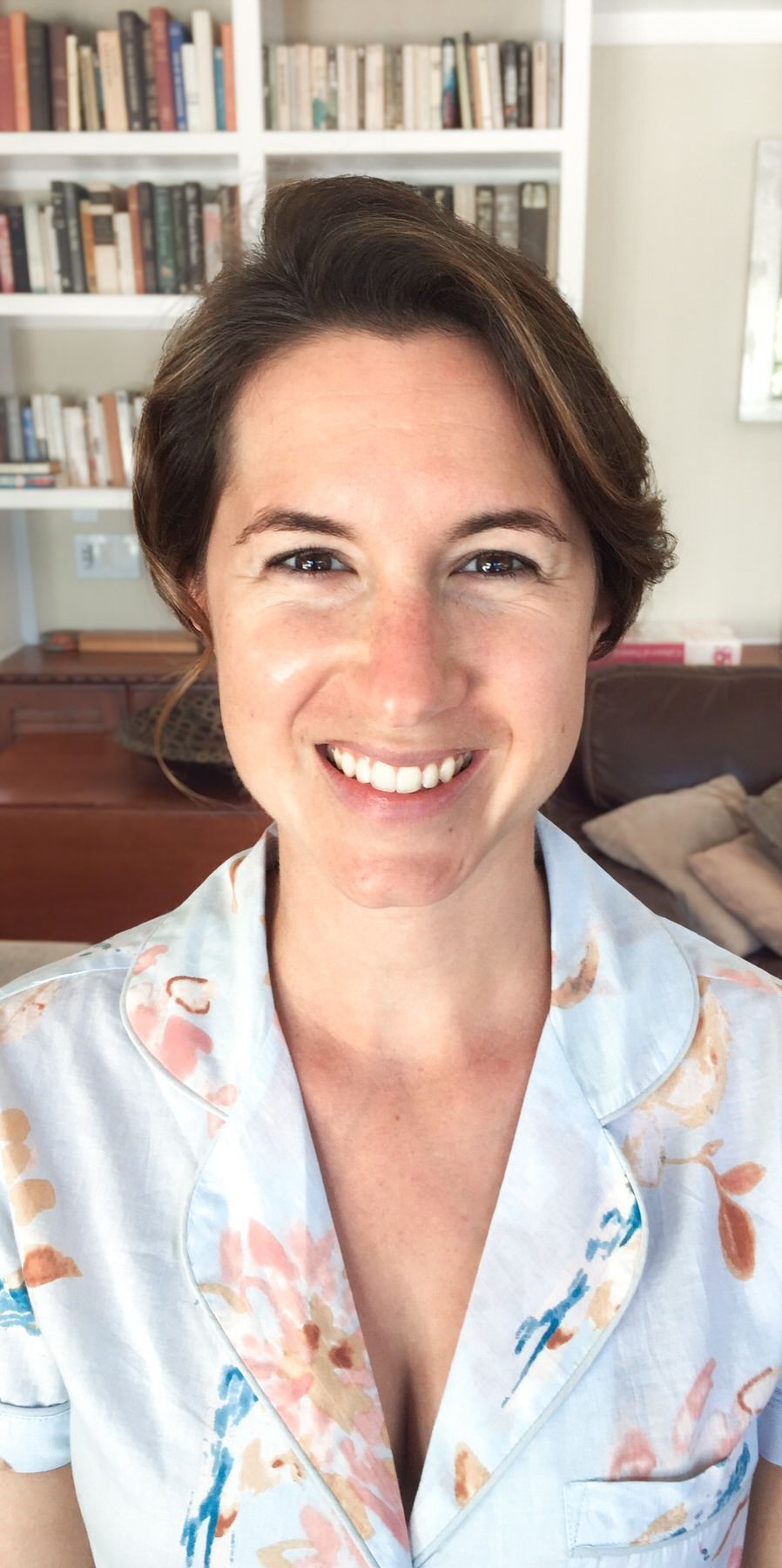 A woman is smiling in front of a bookshelf in a living room. Orange county hair and makeup artist.