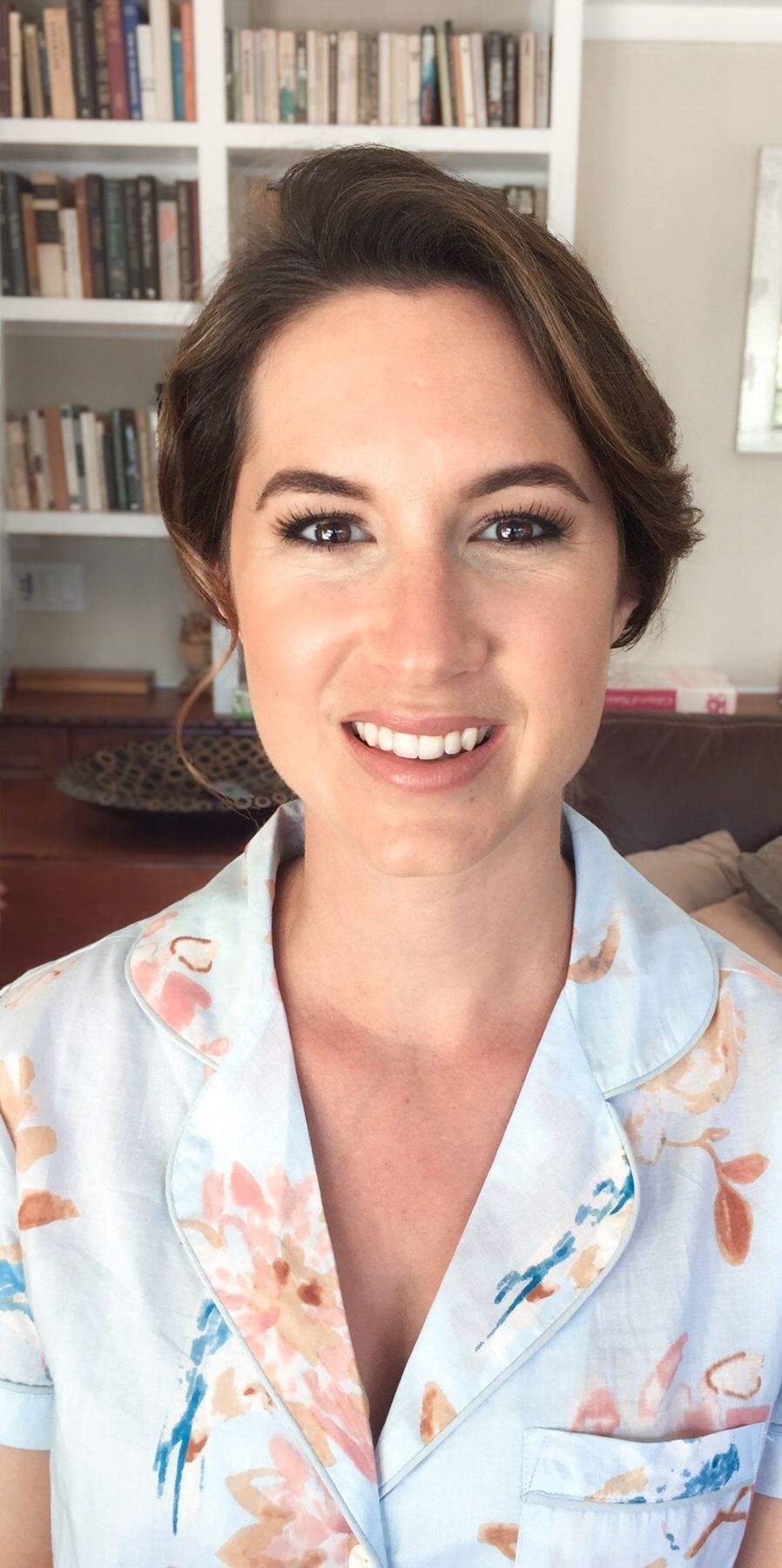 A woman in a floral shirt is smiling in front of a bookshelf. Orange county hair and makeup artist.