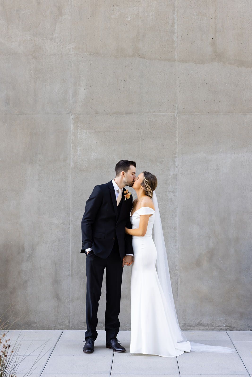 A bride and groom are kissing in front of a concrete wall. Orange county hair and makeup artist.