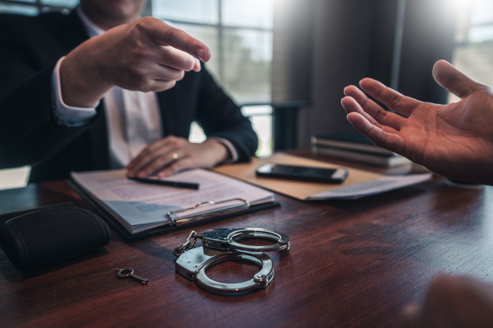Two businesspeople at a desk, one pointing at paperwork beside handcuffs and a phone in a tense meeting