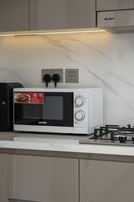 A white microwave sits on a kitchen counter in front of a white marble backsplash, next to a gas stovetop and outlets.