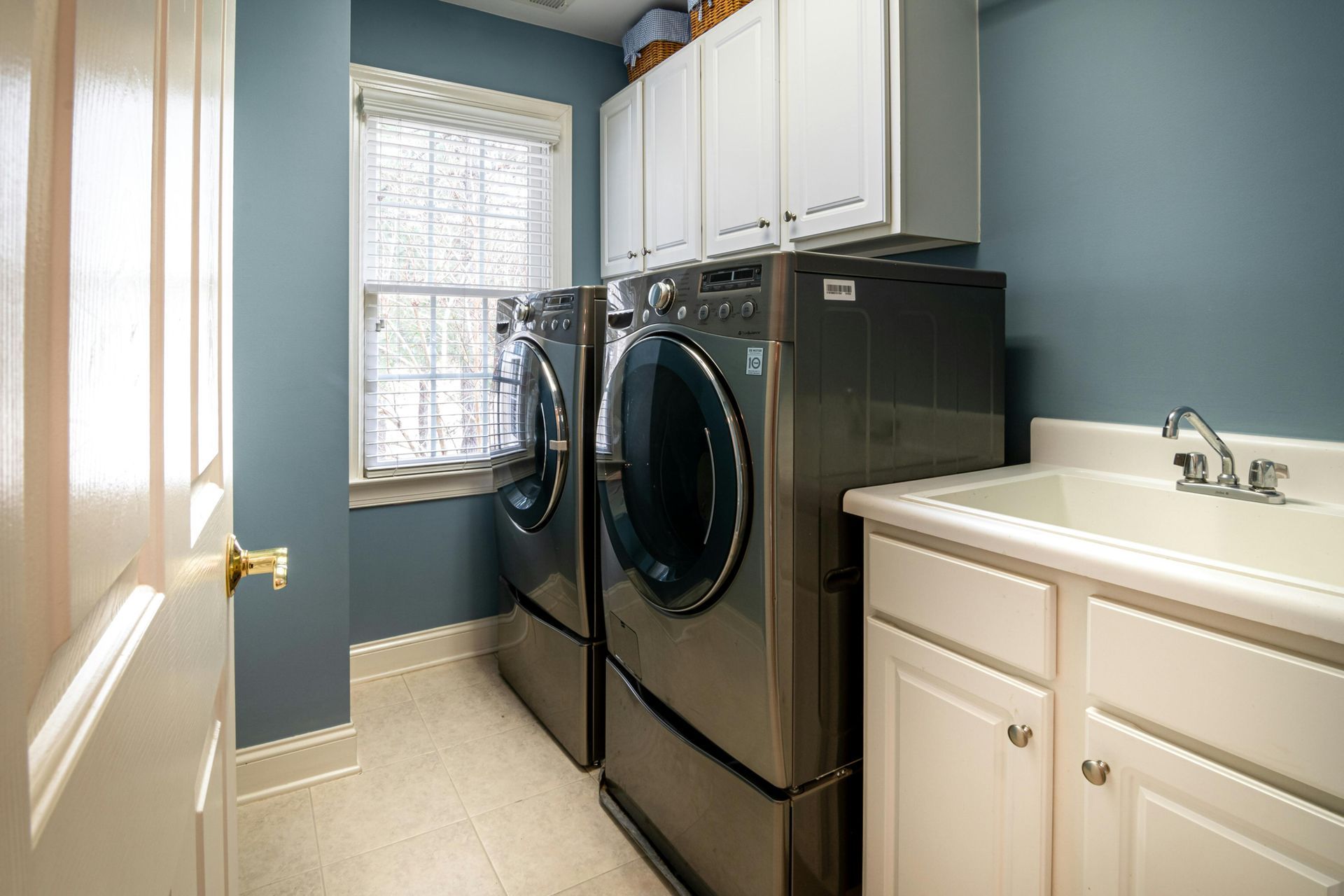 A blue-walled laundry room featuring two dark grey appliances next to a white sink cabinet and a window with blinds.