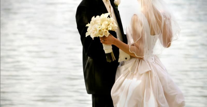 A bride and groom are standing next to each other in front of a body of water.