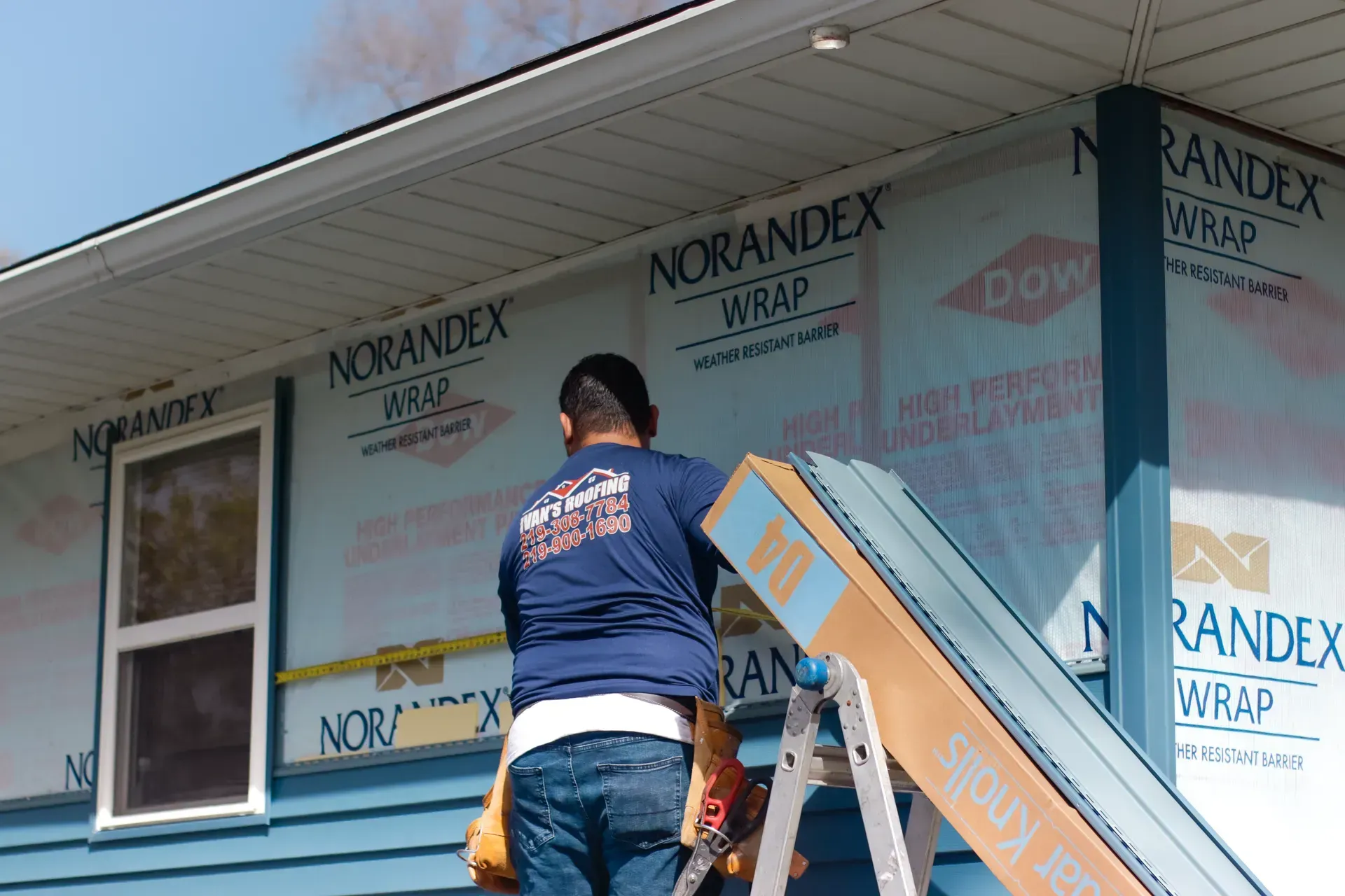 A man is standing on a ladder in front of a house covered in Norandex wrap.