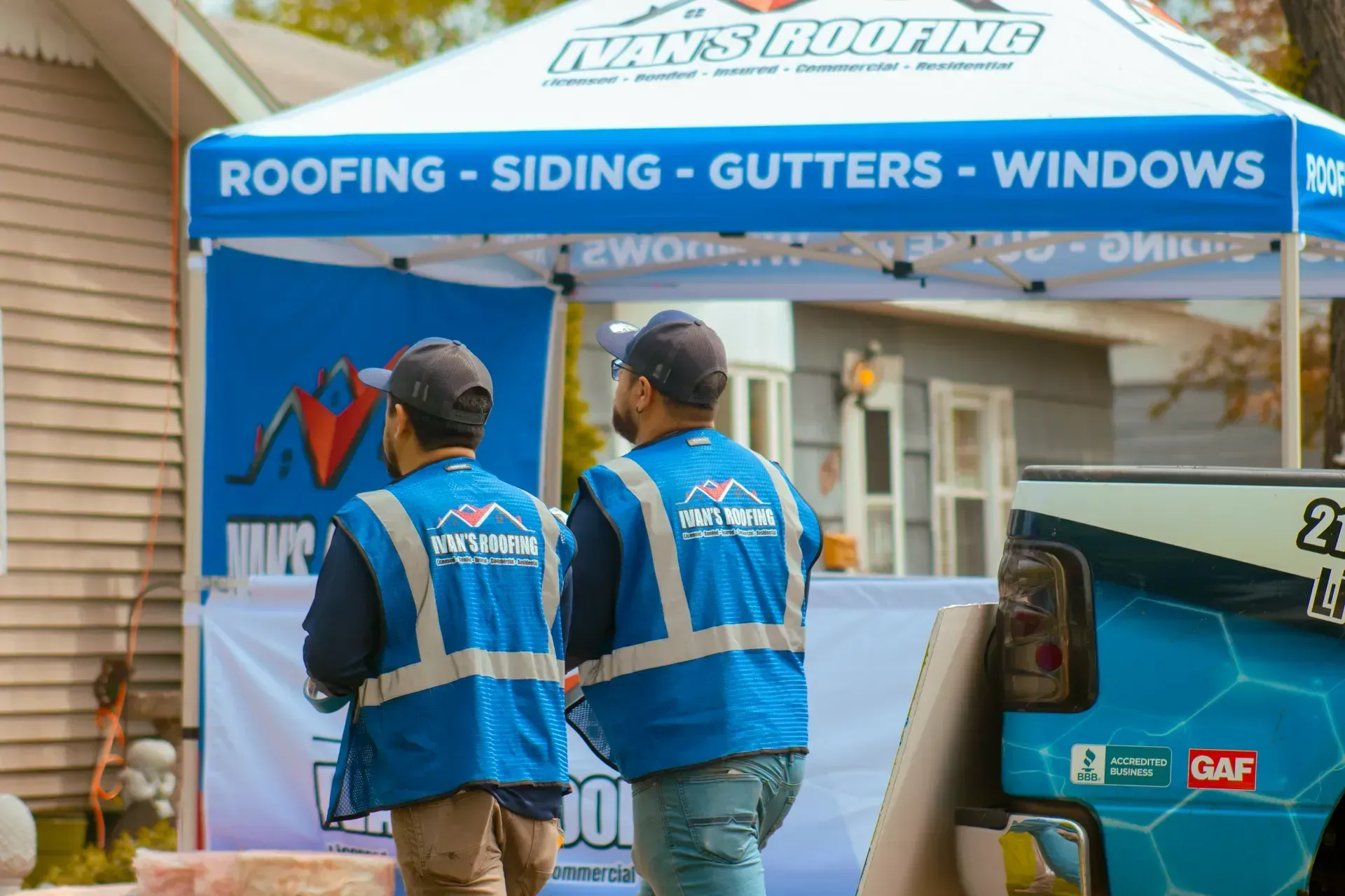 Two men in safety vests are standing in front of a tent that says roofing siding gutters windows.