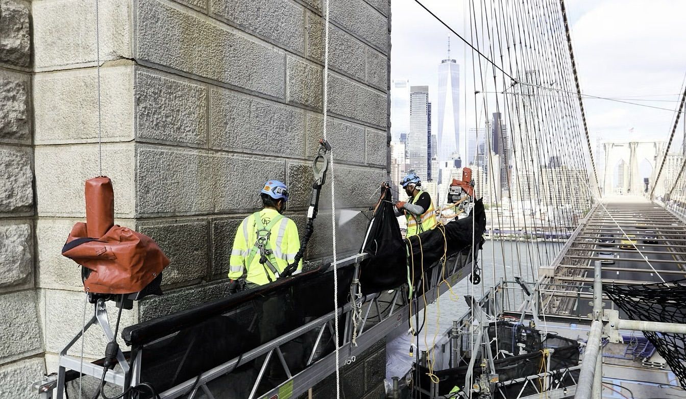 Construction workers on a scaffold, restoring a stone building with city skyline in background.