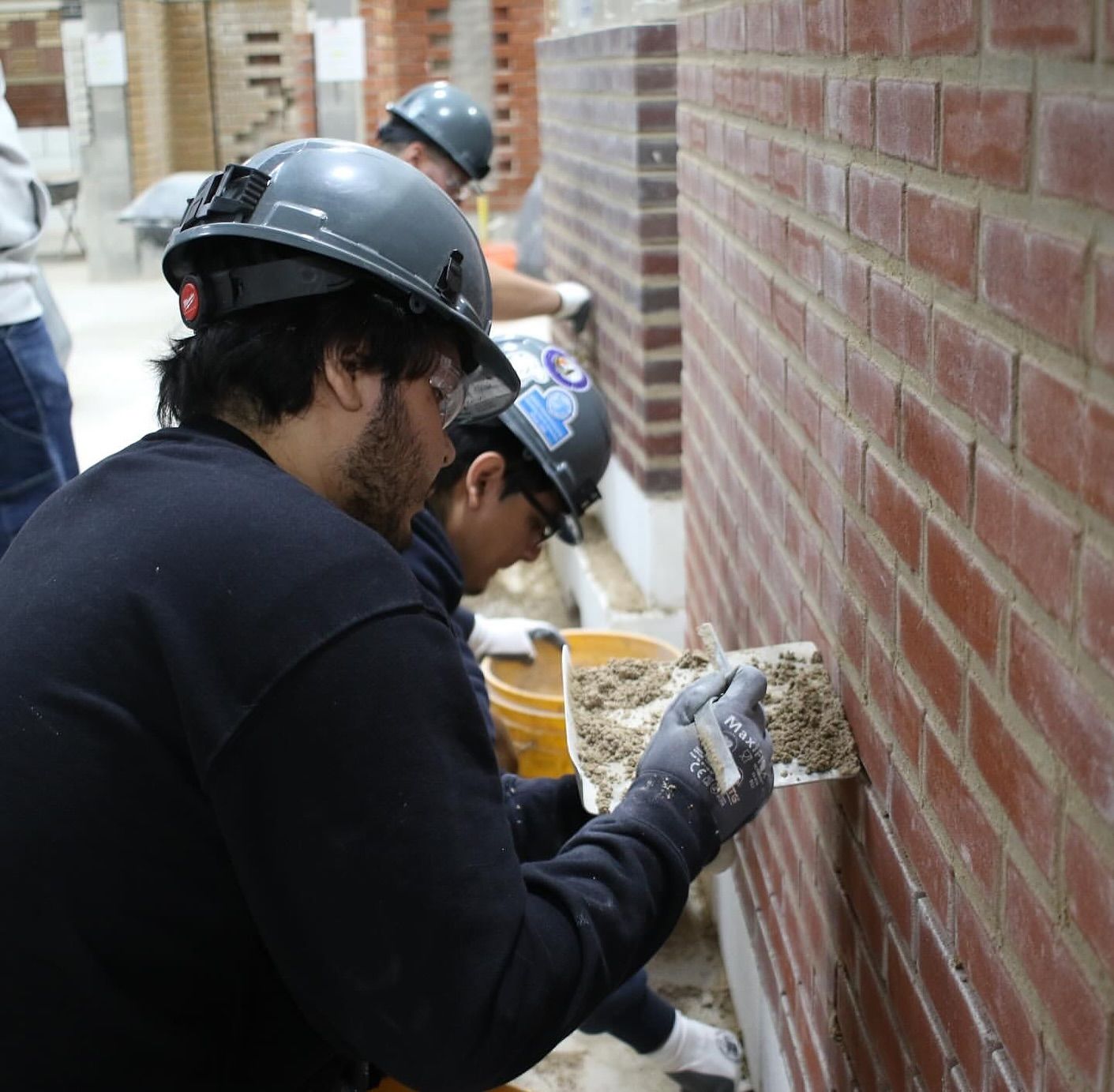 Bricklayers wearing hard hats applying mortar to a brick wall in a construction setting.