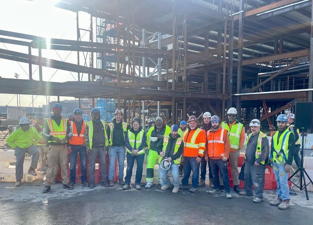 Construction workers pose in front of a steel frame building. Many wear safety vests, outdoors on a sunny day.