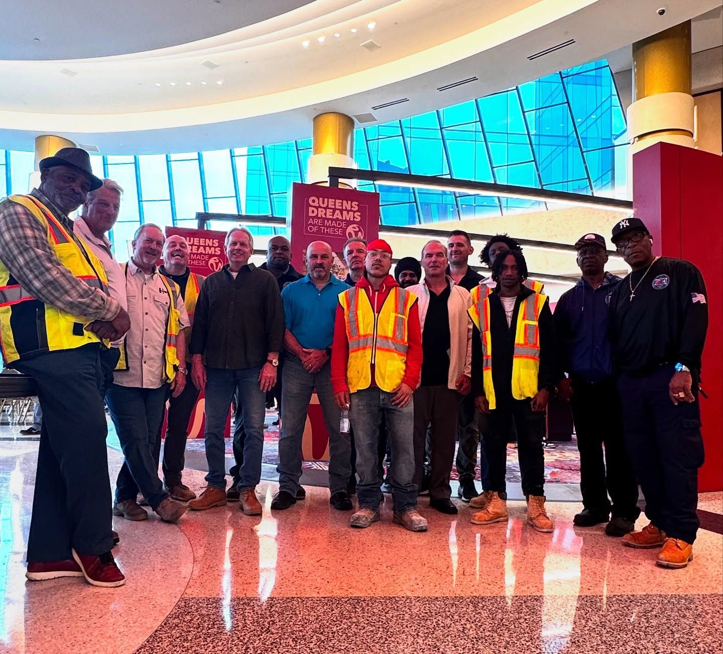 Group of construction workers posing together in a modern building. Many wearing safety vests and work boots.