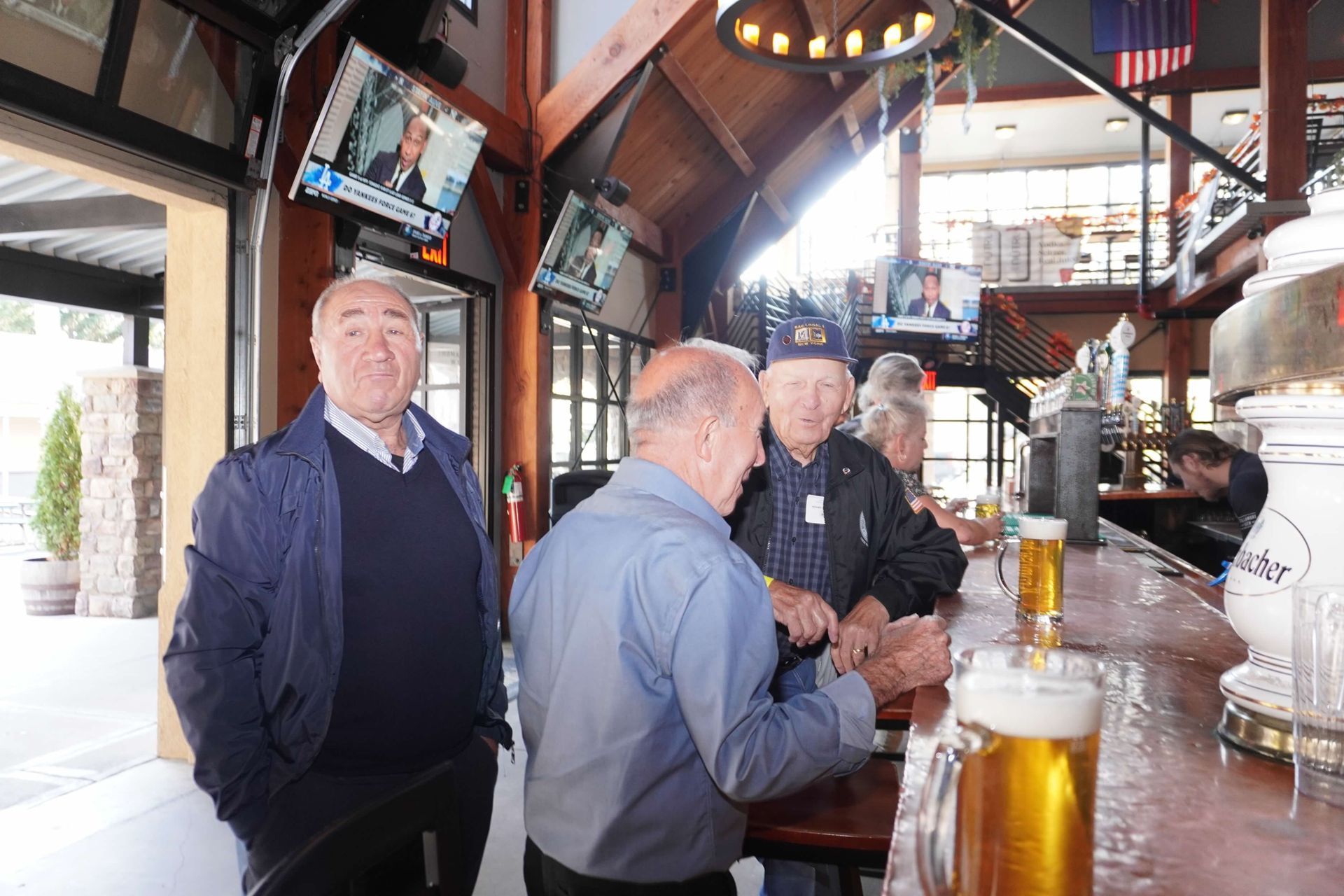 Three men at a bar, two conversing with a beer in front. TVs overhead, open door to the left.