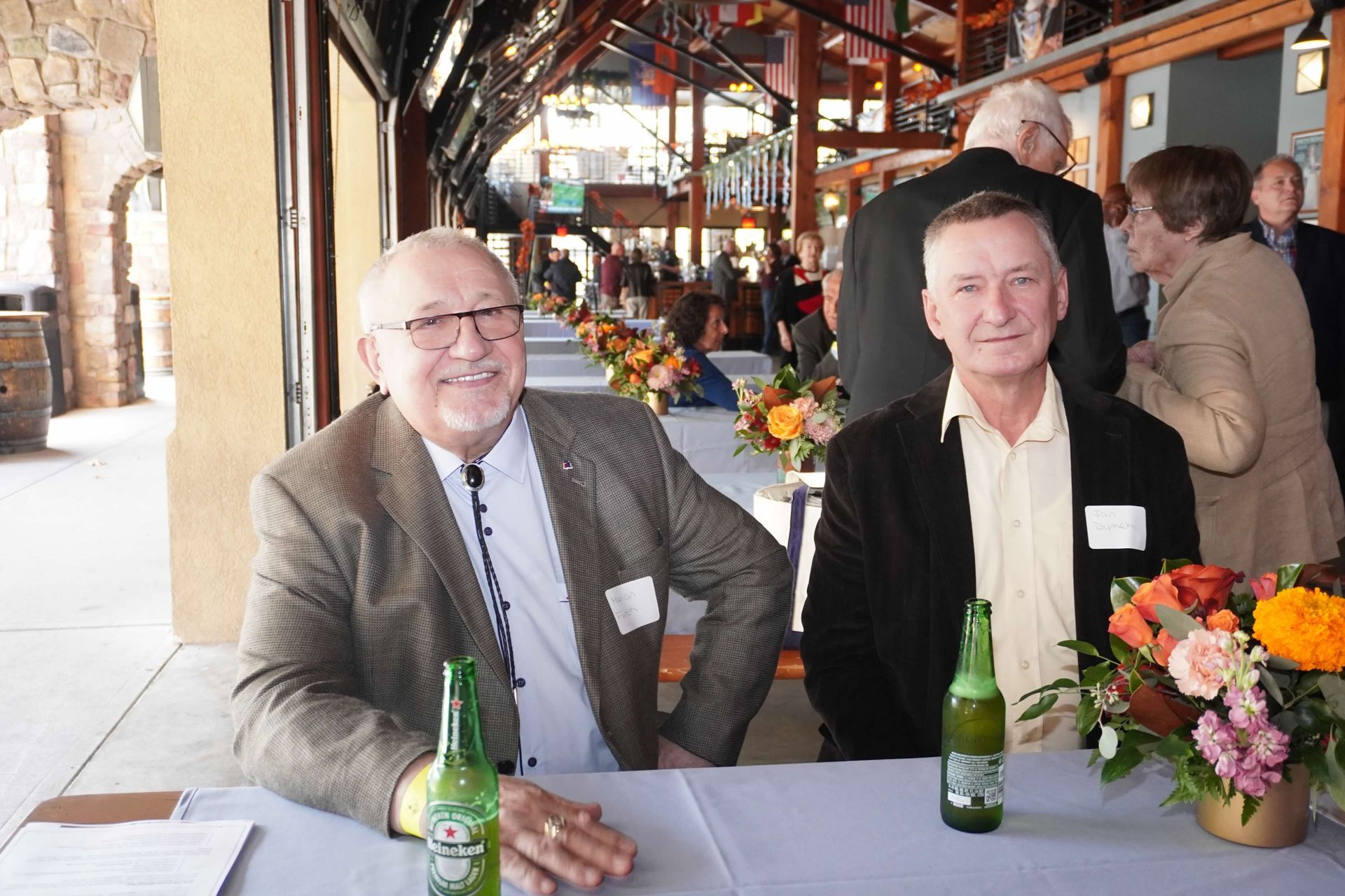 Two men at a table with beer and flowers. They are smiling outdoors at an event.