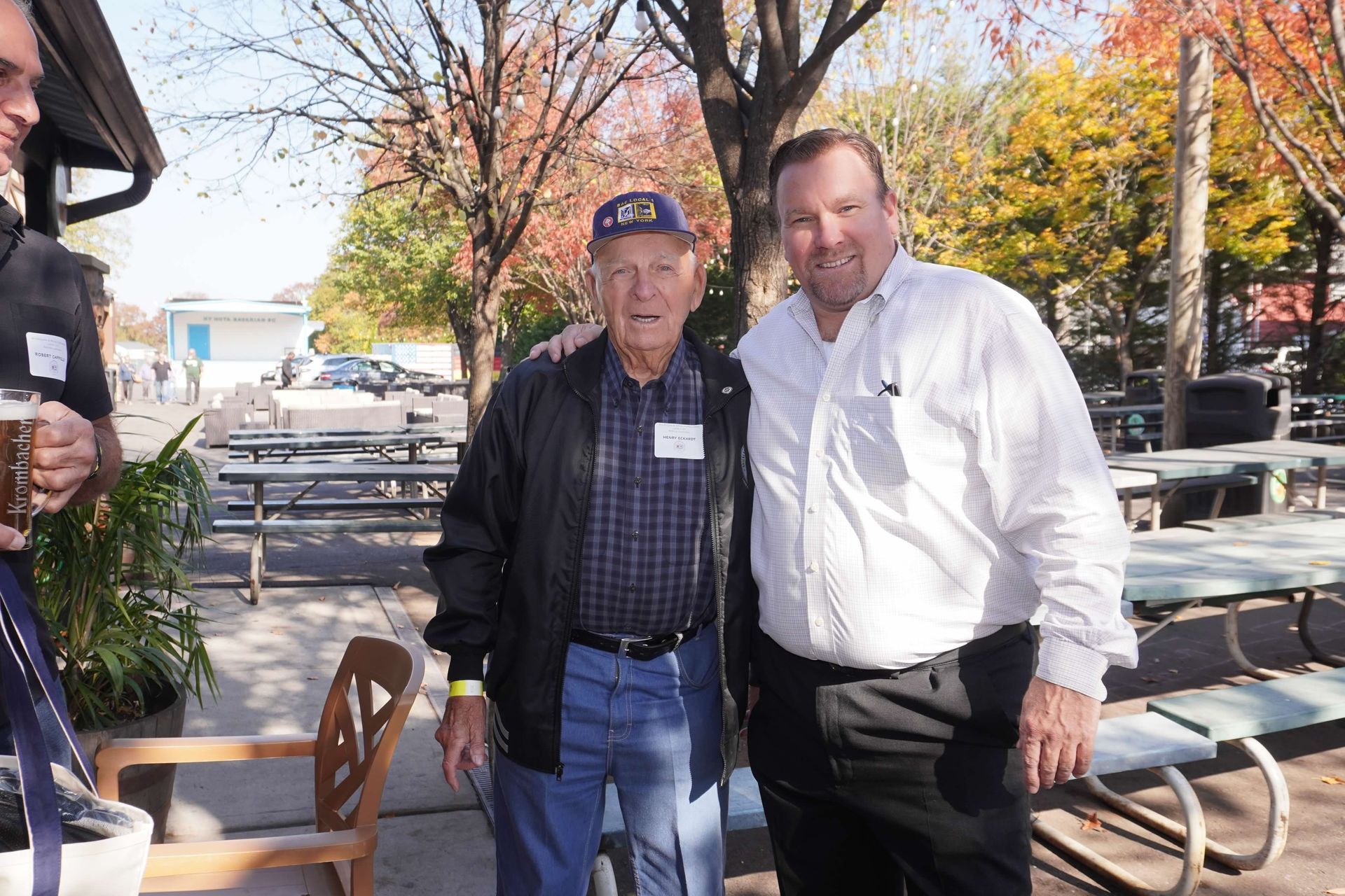 Two men pose outdoors: an older man in jacket and cap with a younger man in a white shirt.