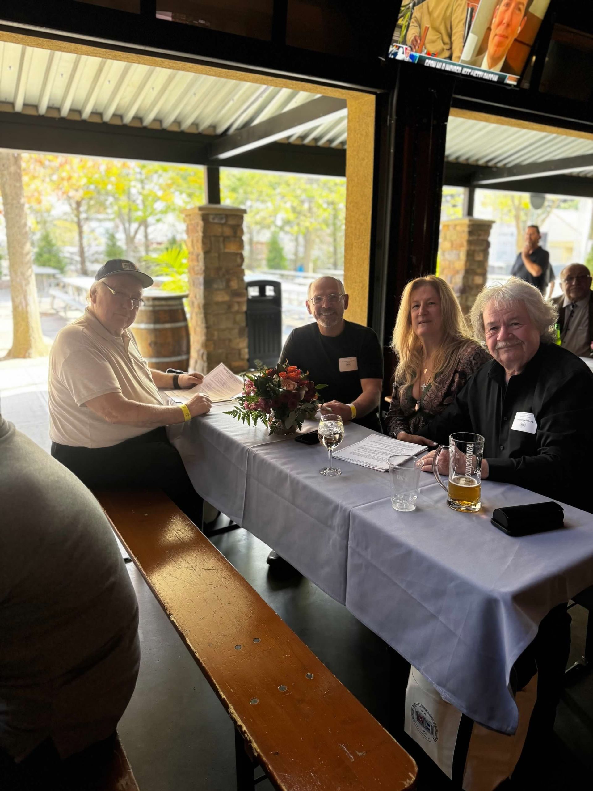 Four people seated at a table in an outdoor dining area, smiling, with drinks and a floral centerpiece.