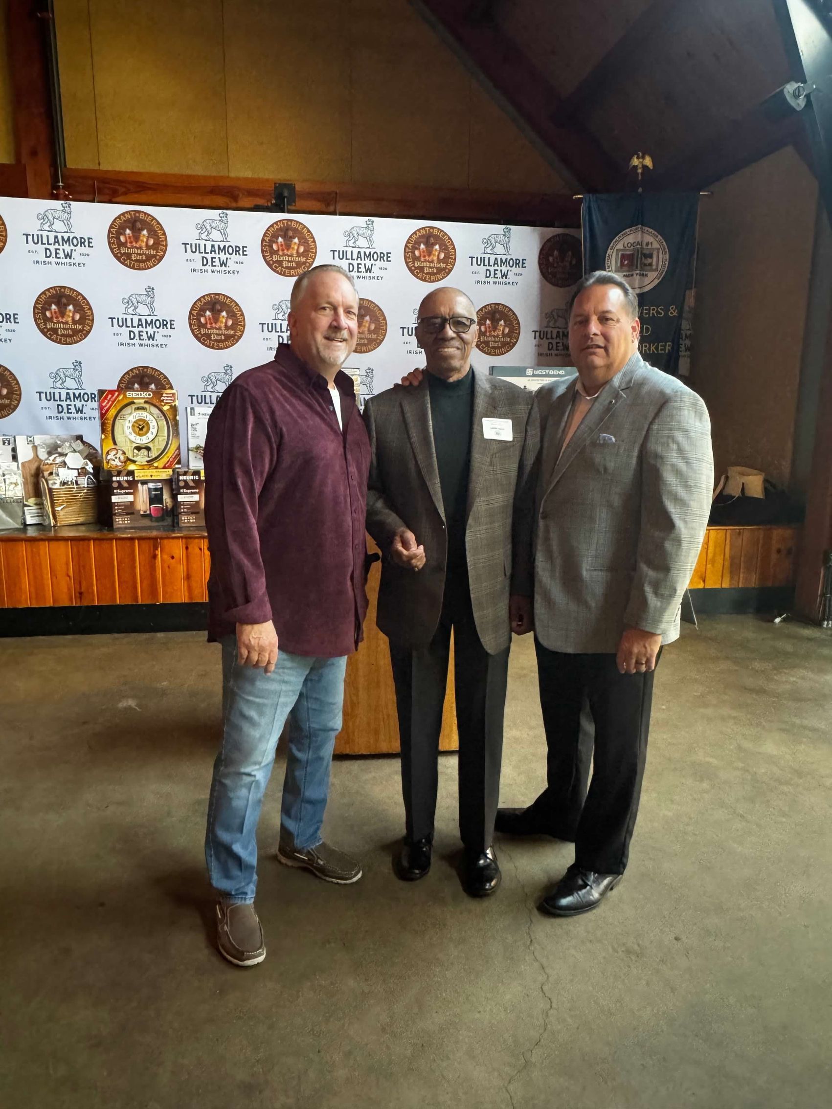 Three men pose for a photo at an event with a logo backdrop. One man is wearing a maroon shirt, another a suit, and the other a gray sport coat.