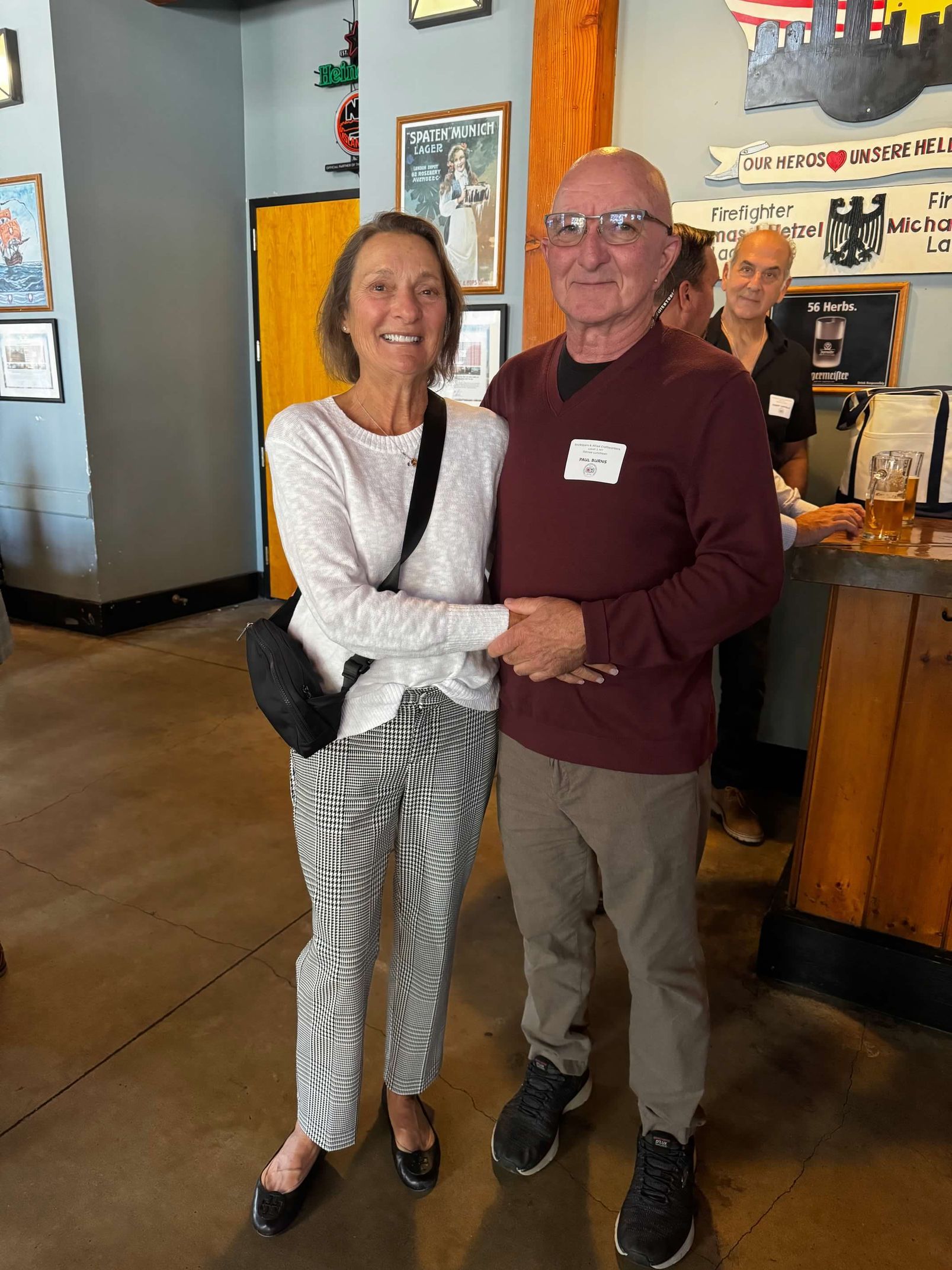 A woman and man hold hands inside a restaurant. The woman wears patterned pants, the man, a maroon shirt.