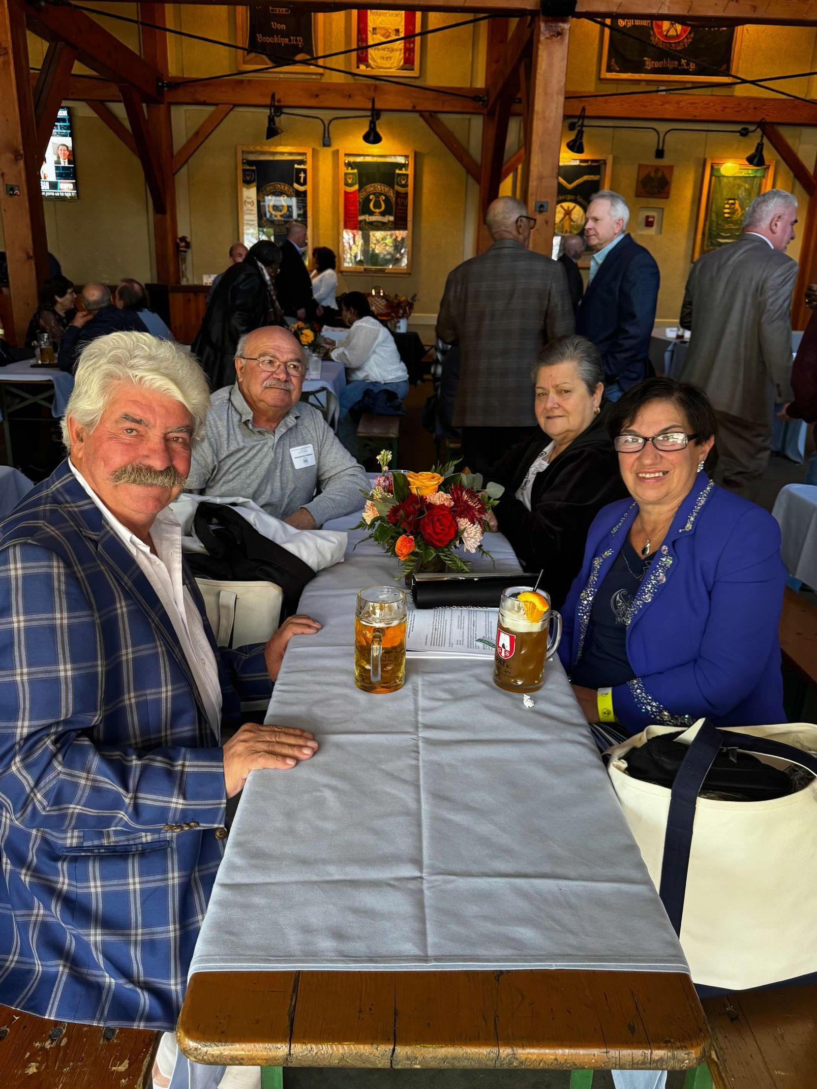 Group of people seated at a table in a beer hall, smiling. Two beers on the table, flower arrangement.
