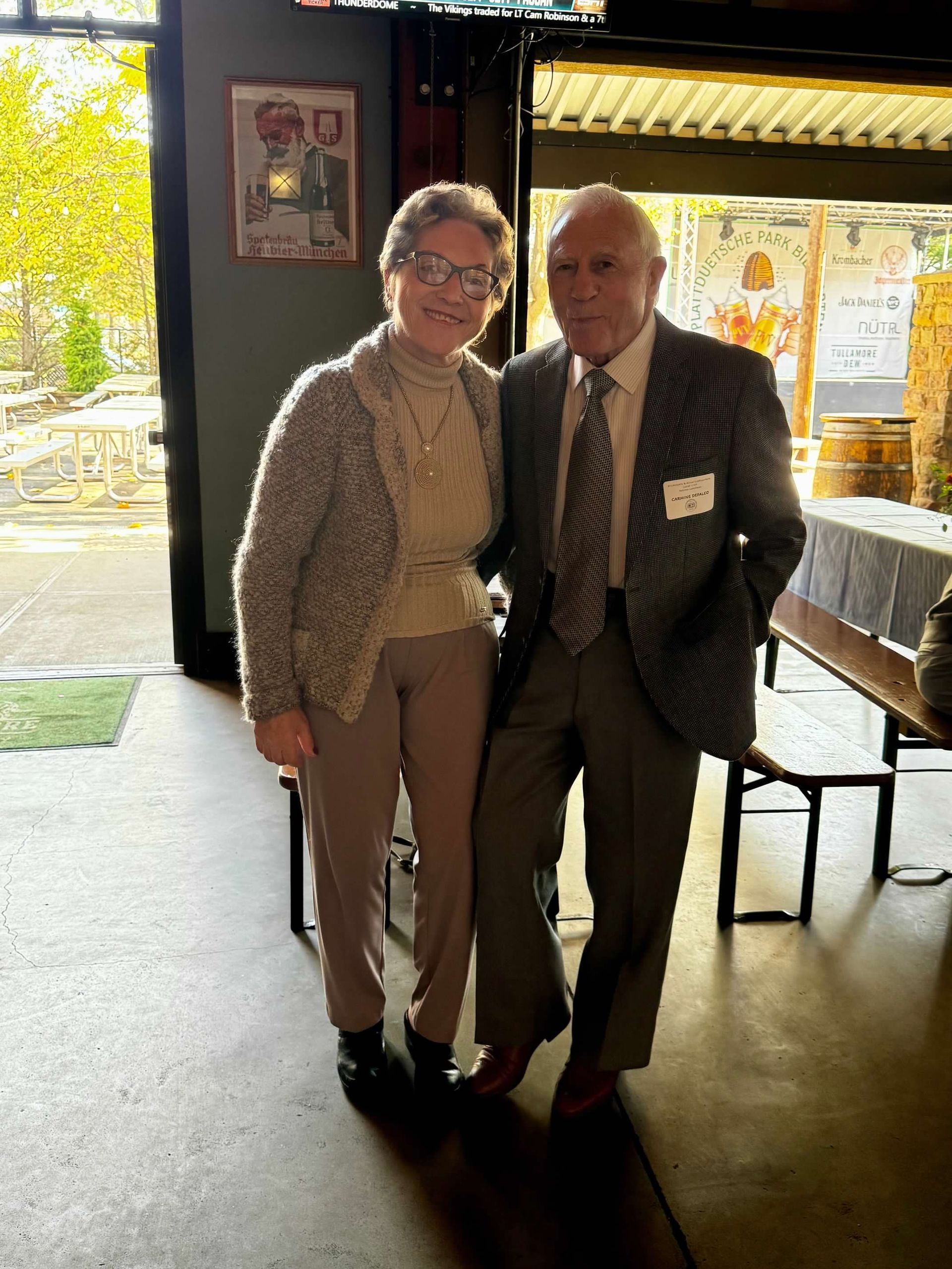 A woman and man stand smiling indoors. The woman wears glasses, a sweater, and pants. The man wears a suit and tie.