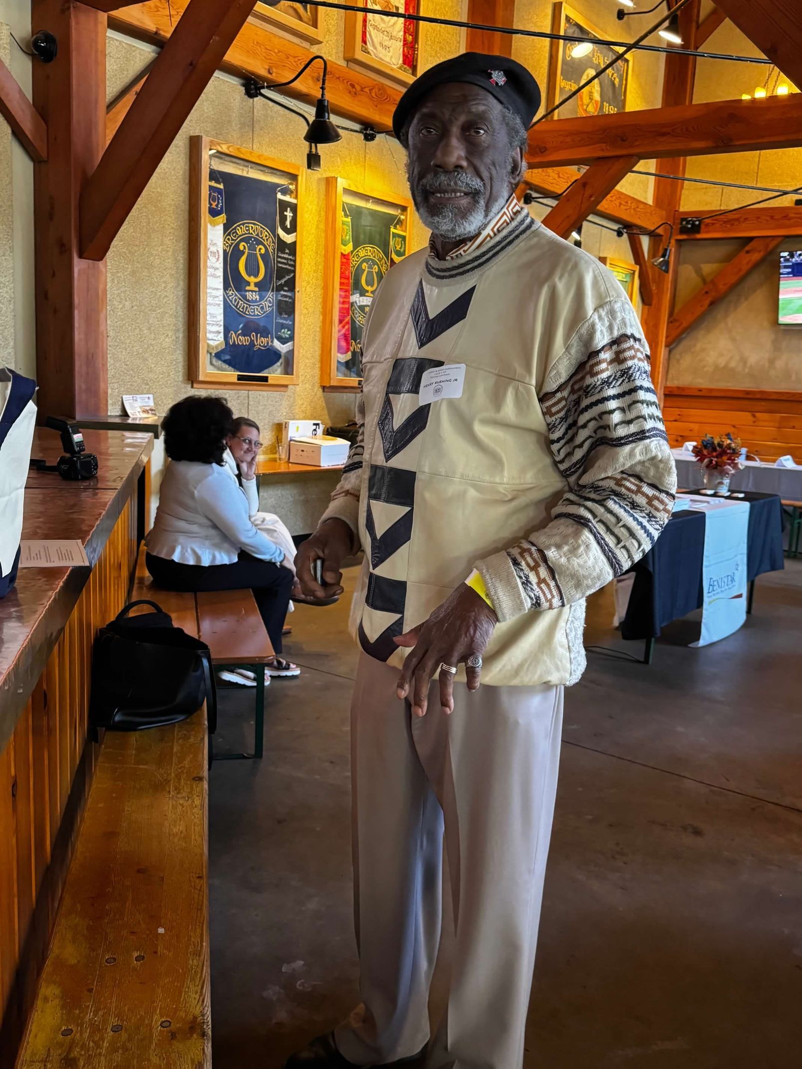 An older Black man in a cream-colored sweater and pants stands indoors; a person sits nearby; decor is in the background.