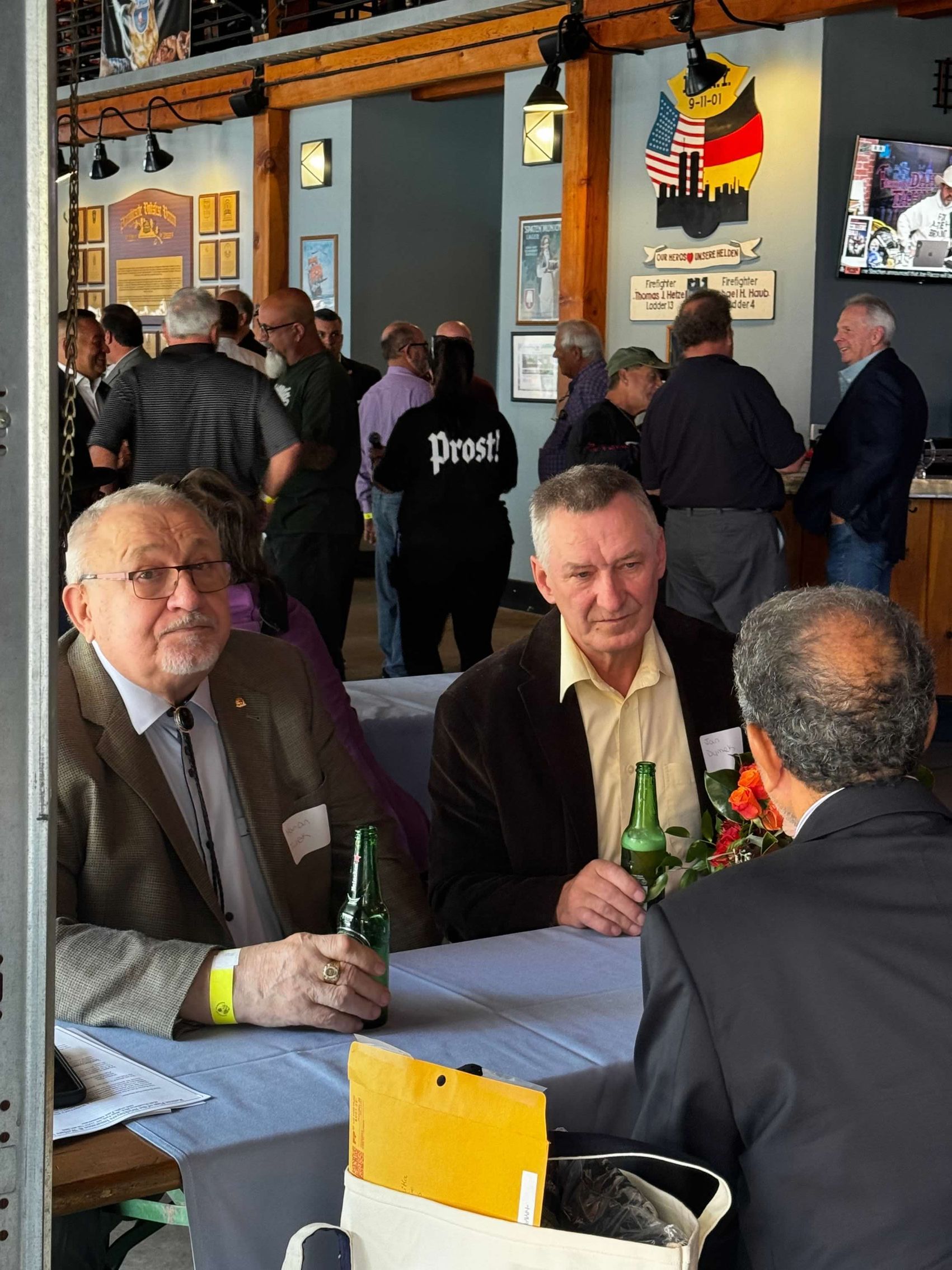 Men at a table in a restaurant, talking and drinking beer, with a crowd in the background.