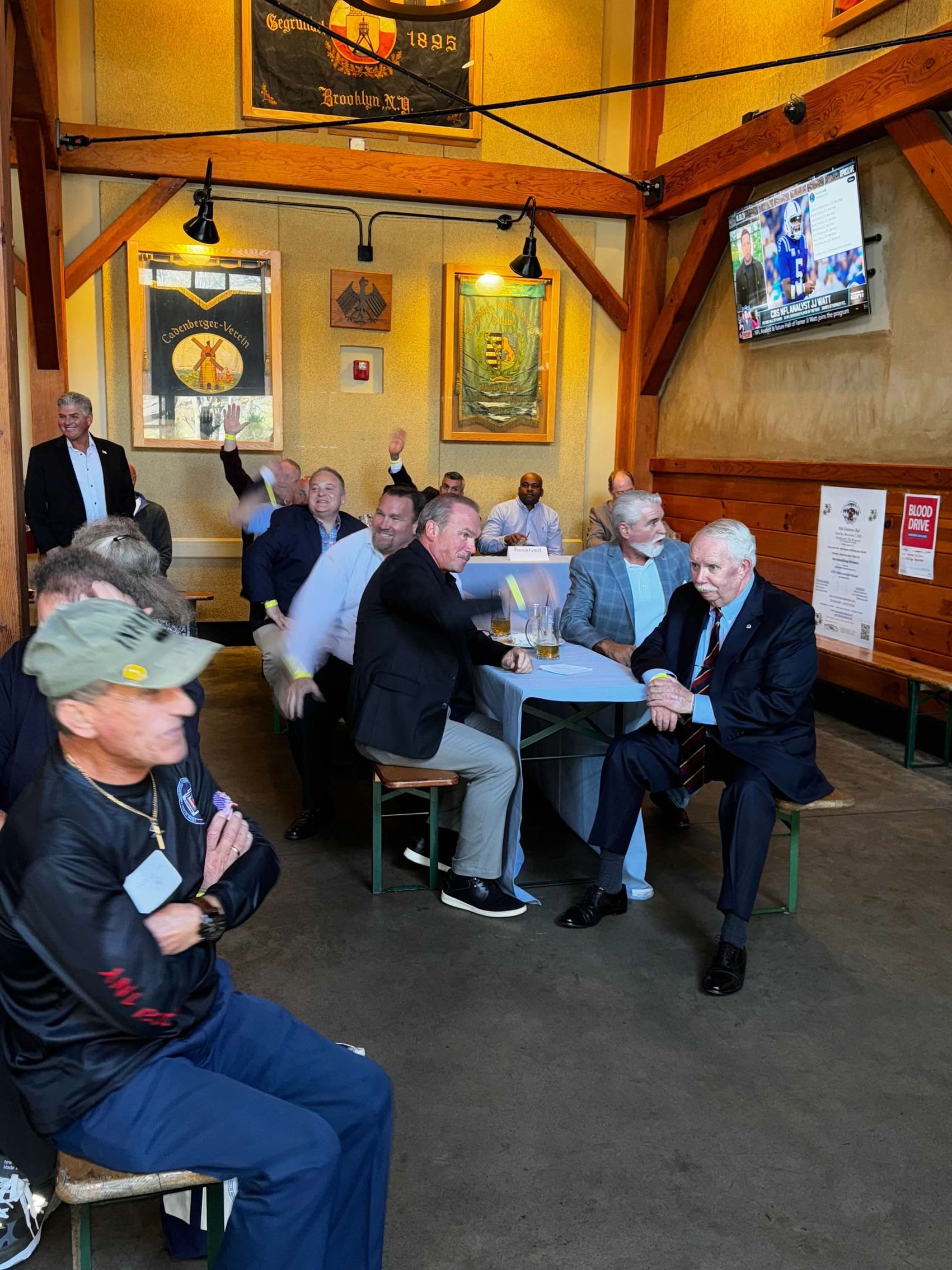 People in a German beer hall; some seated, some standing. Men watching a TV, one man raises his arm.