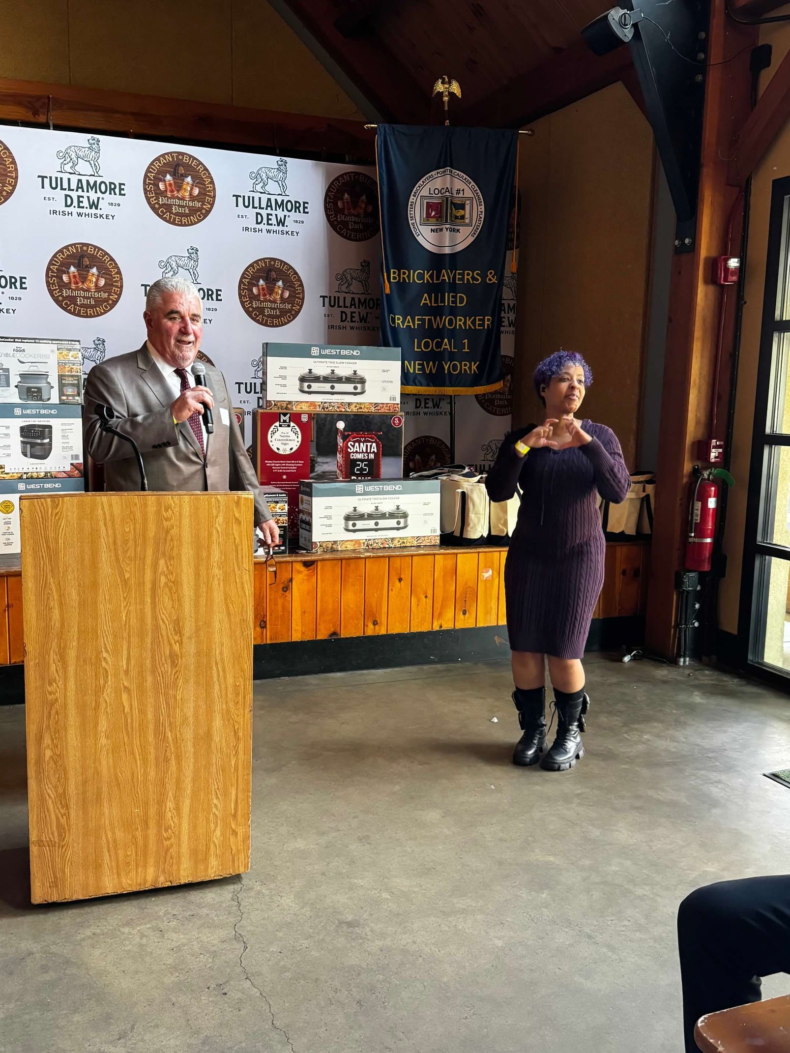 Man at podium, woman signing, event with banners, tables with items.