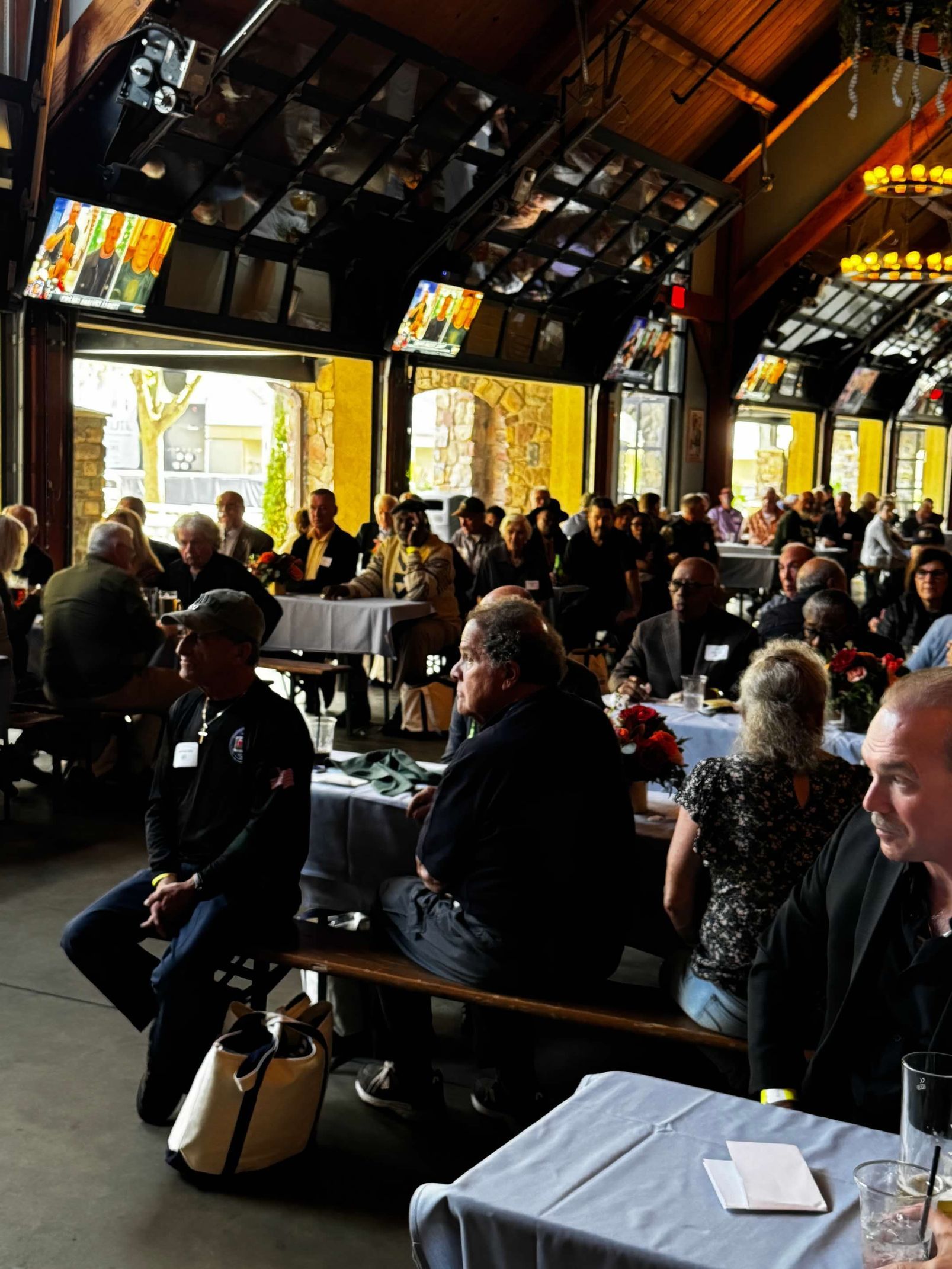 Large group of people at an event, seated at tables in a building with glass windows and an exposed wooden ceiling.