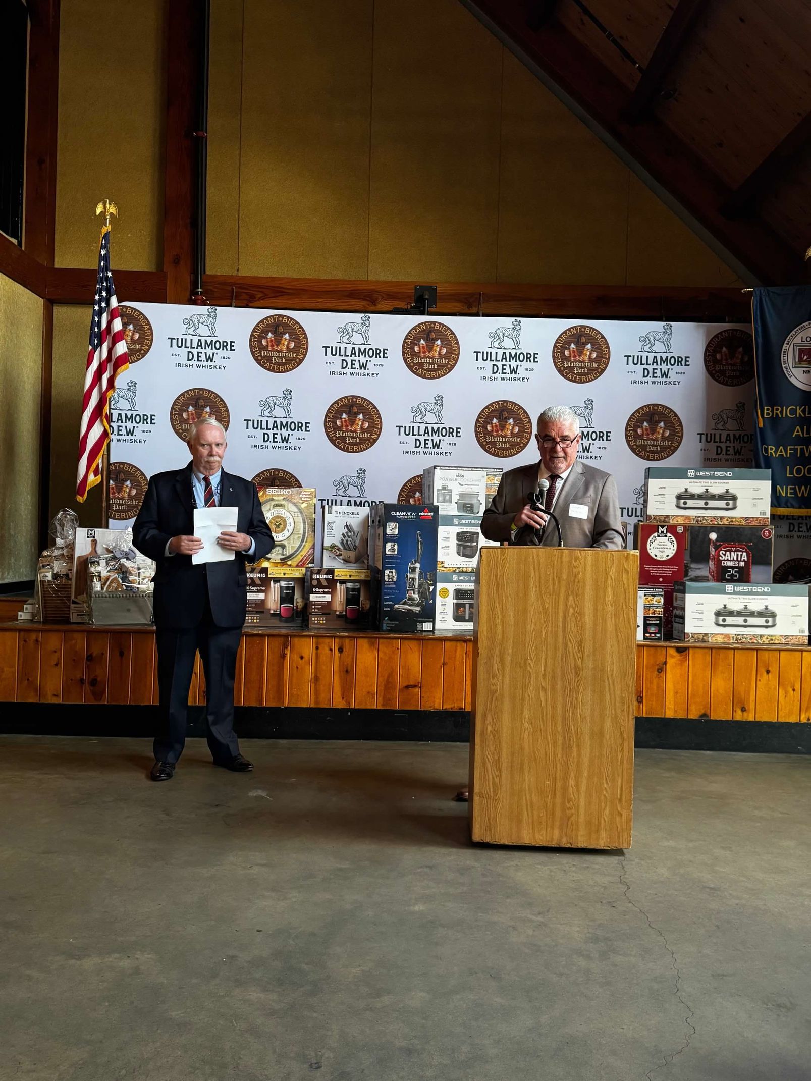 Two men at podium. One speaks, other stands with notes. American flag in the background.