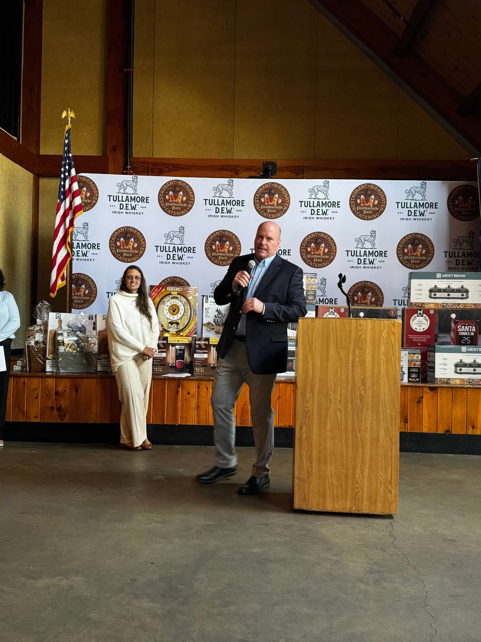 Man speaking at podium, woman standing beside him. Event backdrop, American flag.