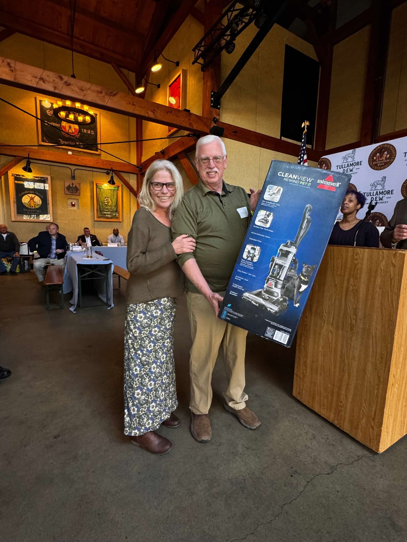 Man and woman hold a framed print indoors; woman smiles, man wears a pin and beige pants.