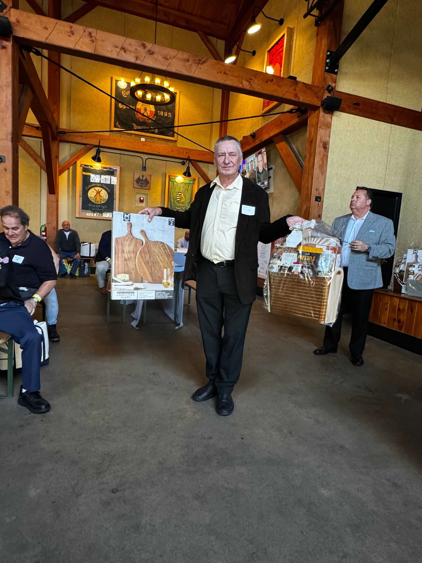 Man in suit holds a framed map and gift basket at an indoor event, other people seated.