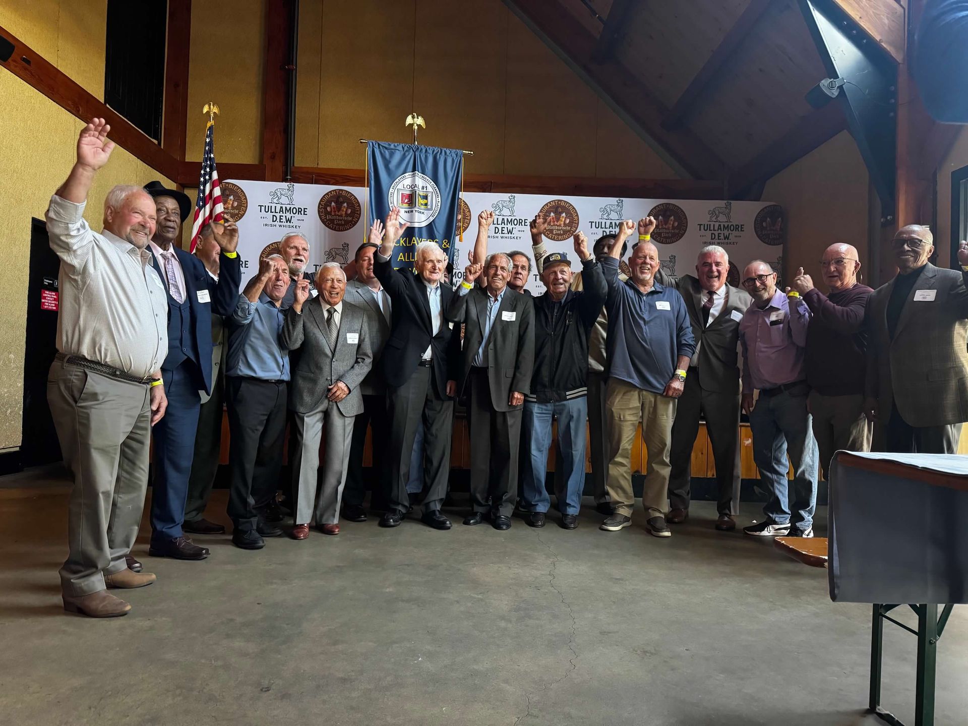 Group of men indoors, some raising hands, with a banner and American flag.