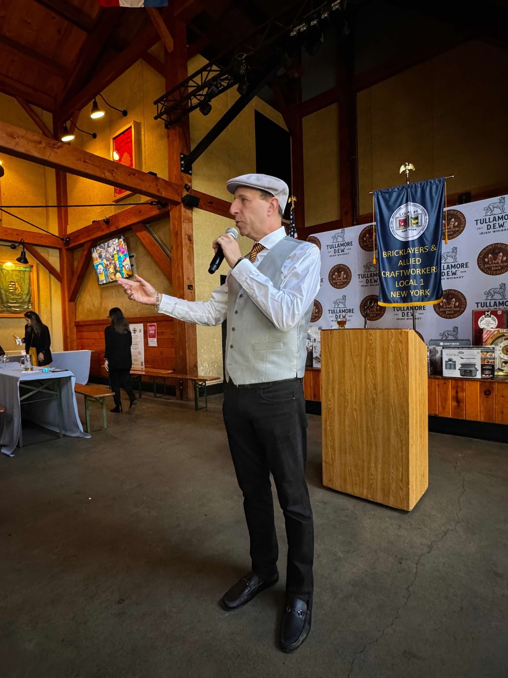Man in vest and hat speaks at podium in a hall. He holds item, gesturing.
