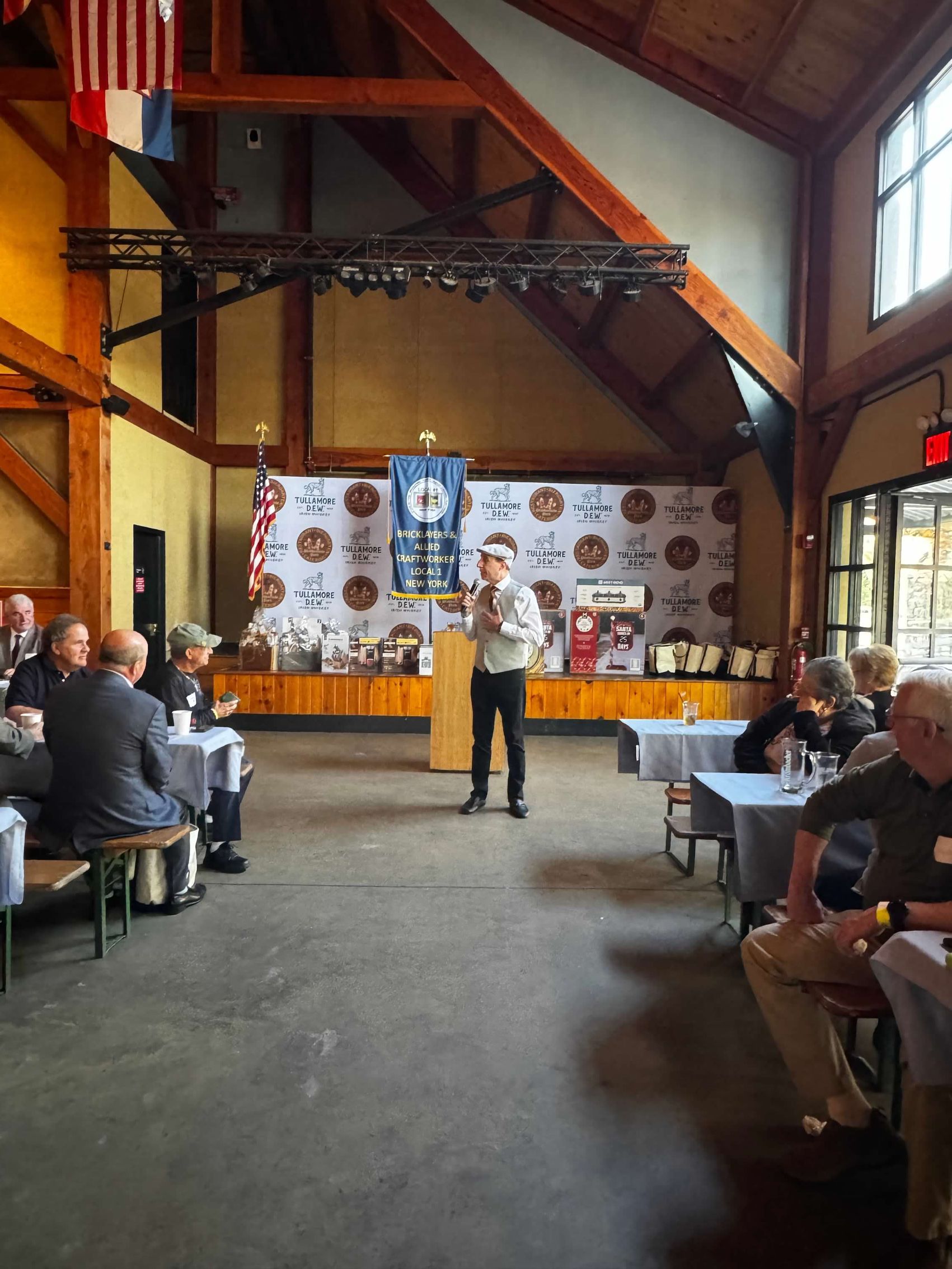Man speaking at a podium in a lodge-like hall, audience seated at tables, awards backdrop.