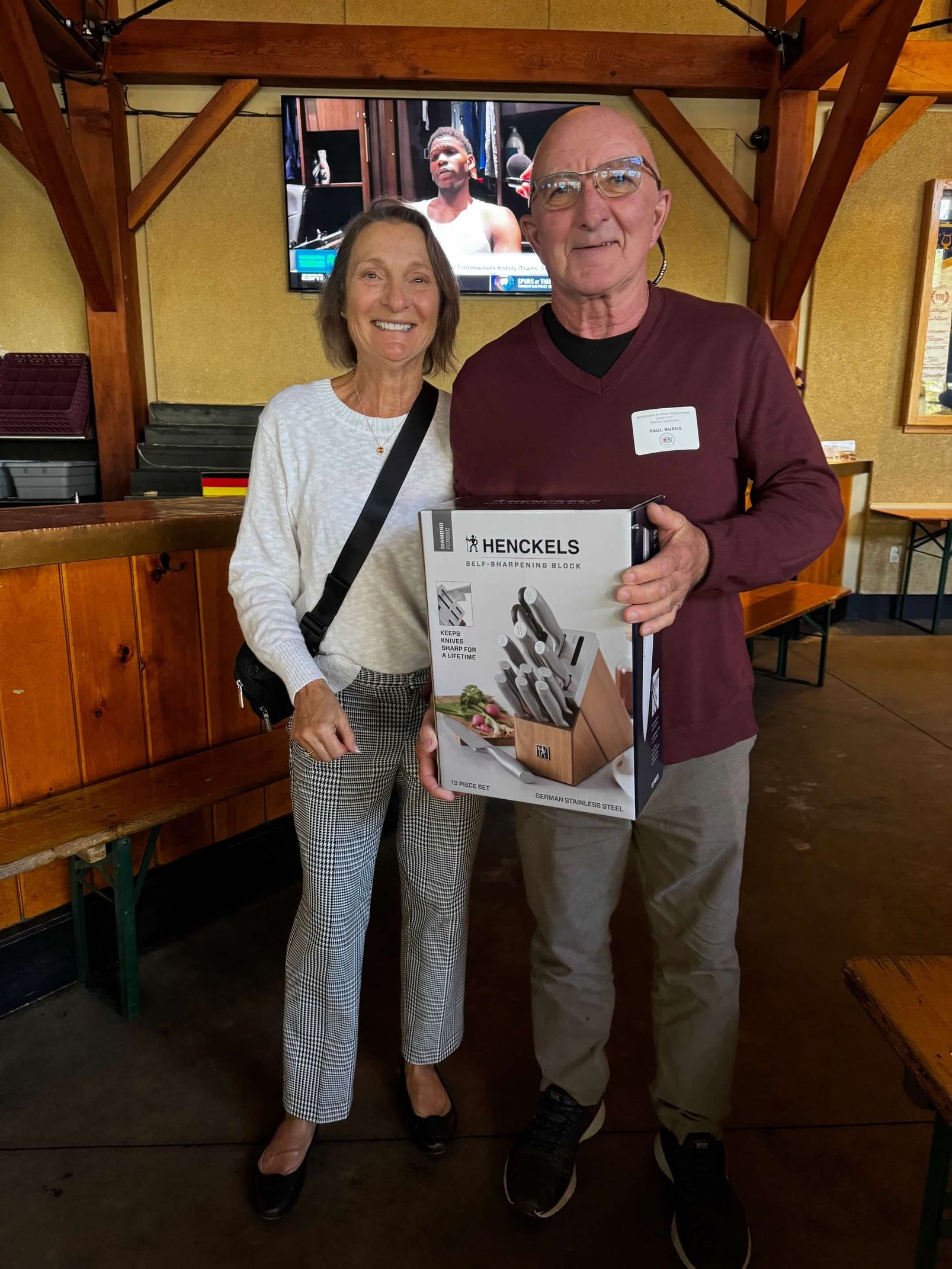 Woman and man pose indoors, man holding a magazine, both smiling. Wooden interior, TV in background.