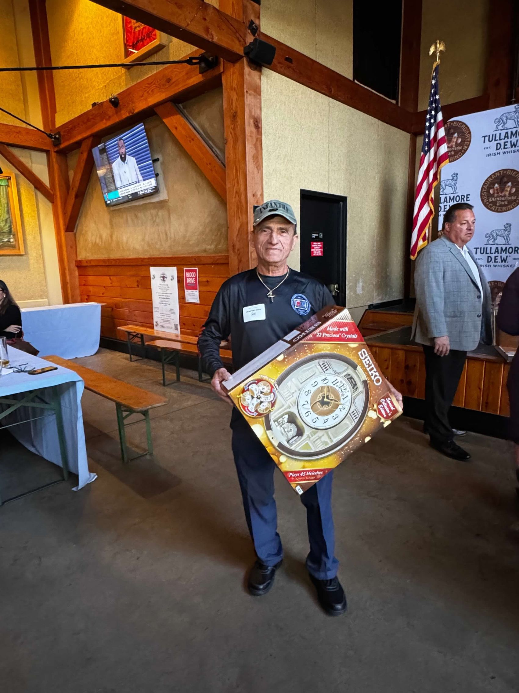 Man in uniform holds prize box at an event, inside a building with wooden beams and an American flag.