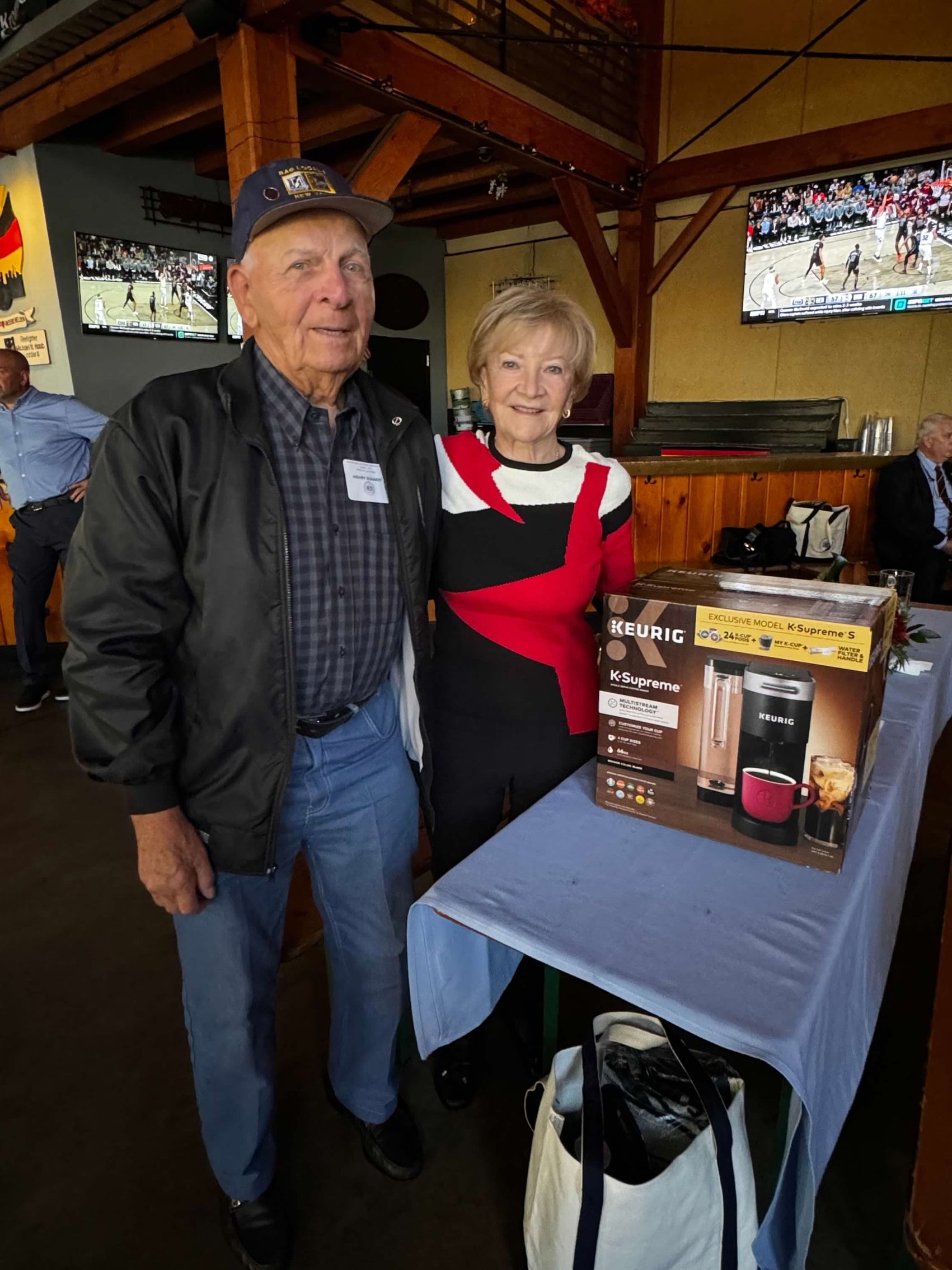 An elderly couple poses with a boxed coffee maker at an event in a restaurant setting.