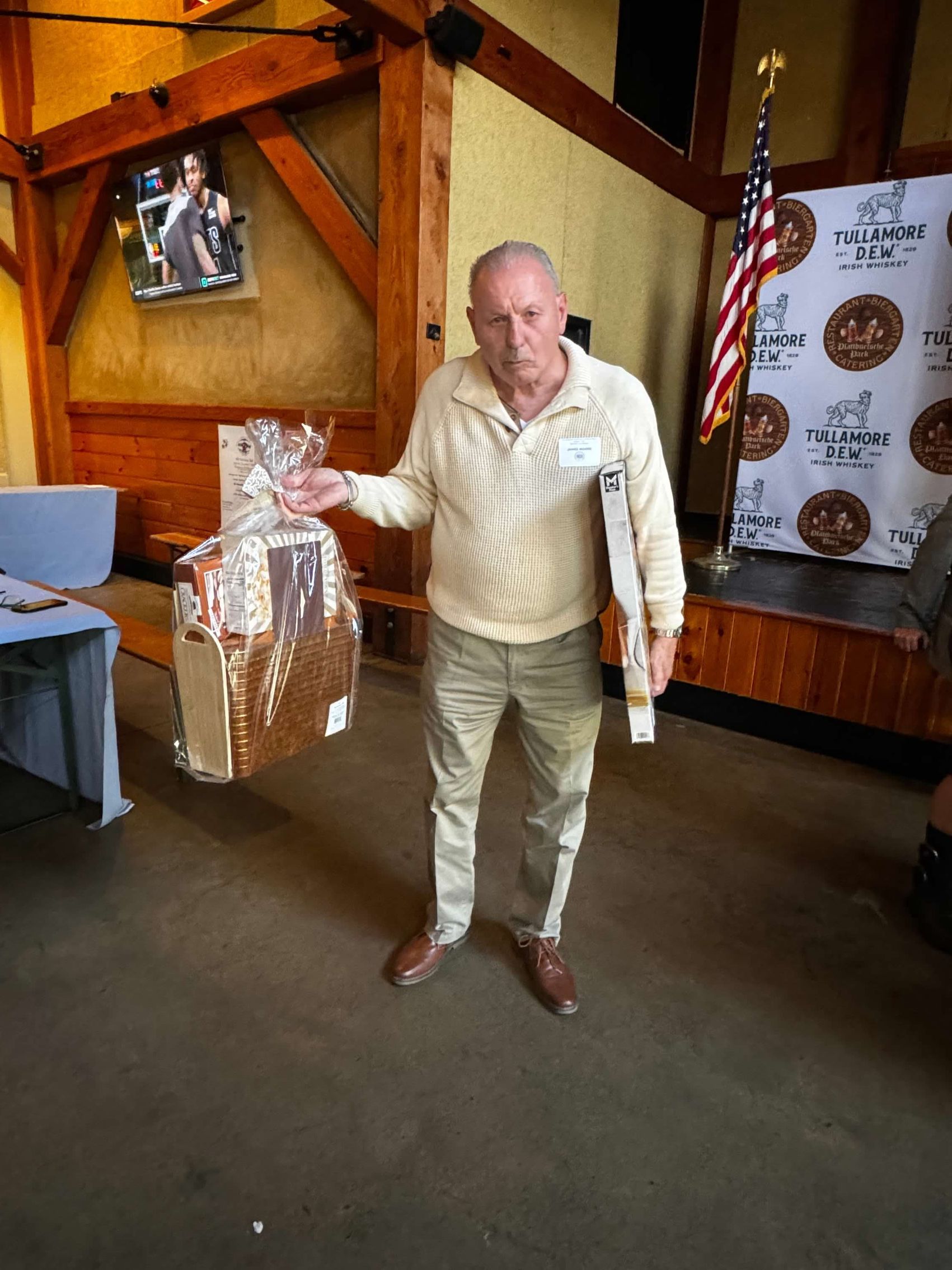 Man in beige sweater and khaki pants holds gift basket and plaque at an event.