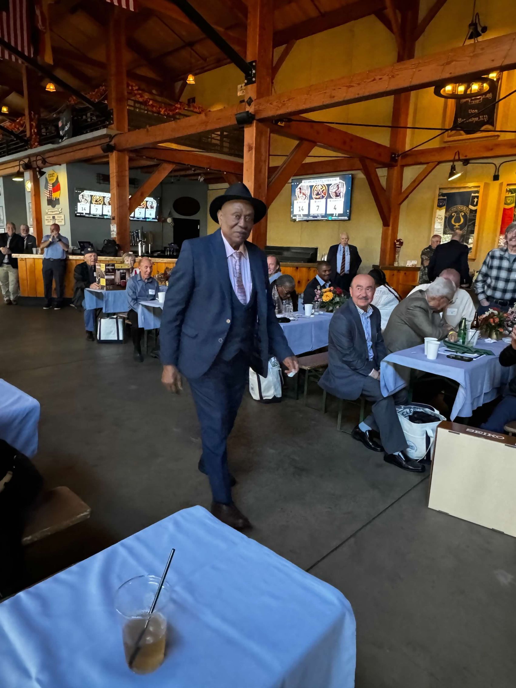 Man in blue suit and hat walks through a room with tables set for a gathering.