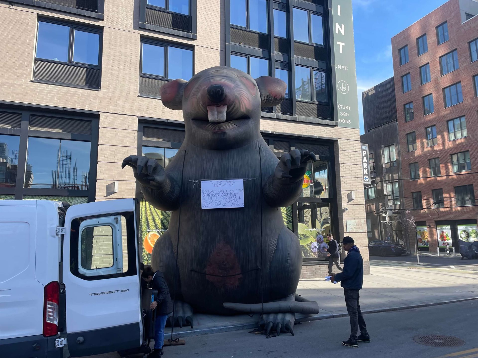 Giant inflatable rat stands outside a building, two people nearby; a videographer and person next to a van.