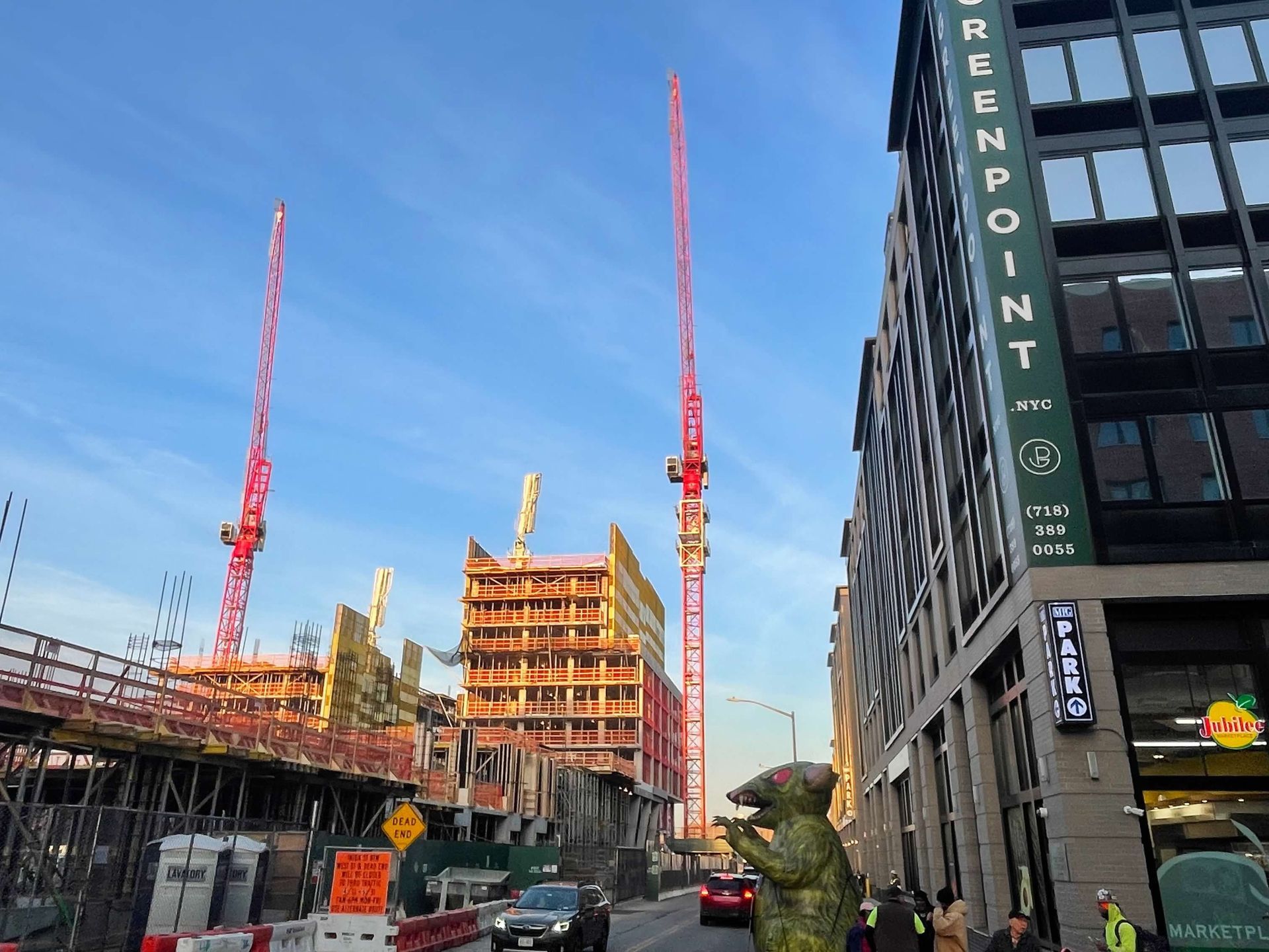 Construction site with two red cranes, building facade, and a street with a dinosaur statue.