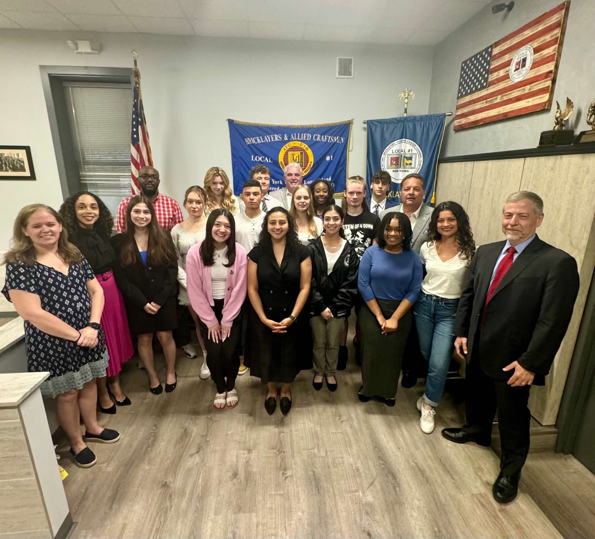 Group of people posing in a room with flags, smiling. A man in suit stands at the right.