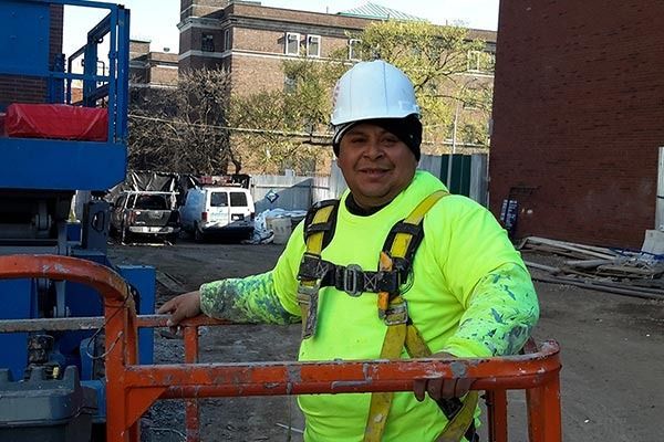 Construction worker in neon yellow shirt, white hard hat, and safety harness, on a lift, smiling outside.